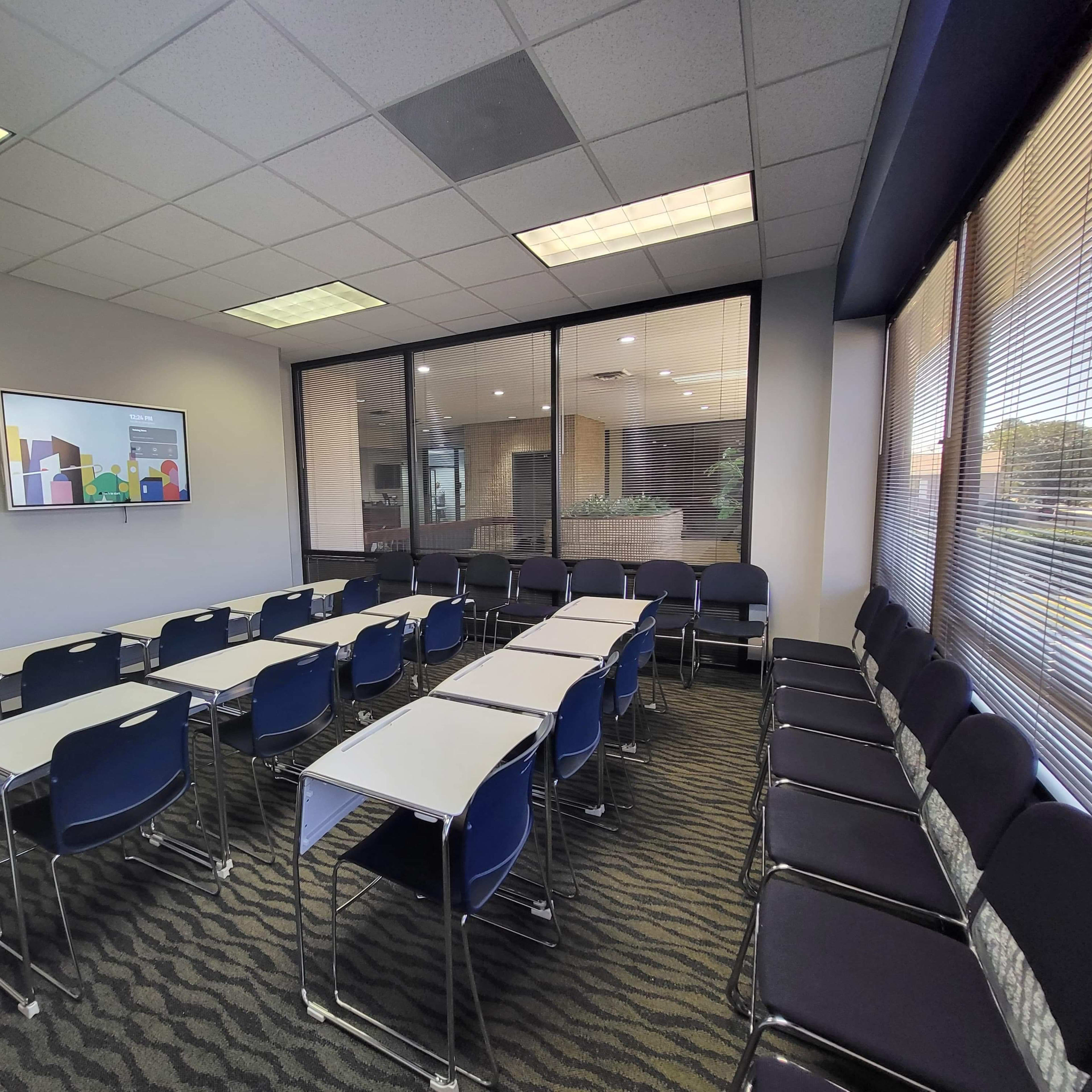 A conference room features rows of chairs and desks facing a wall-mounted screen, with large windows providing a view of the outside.