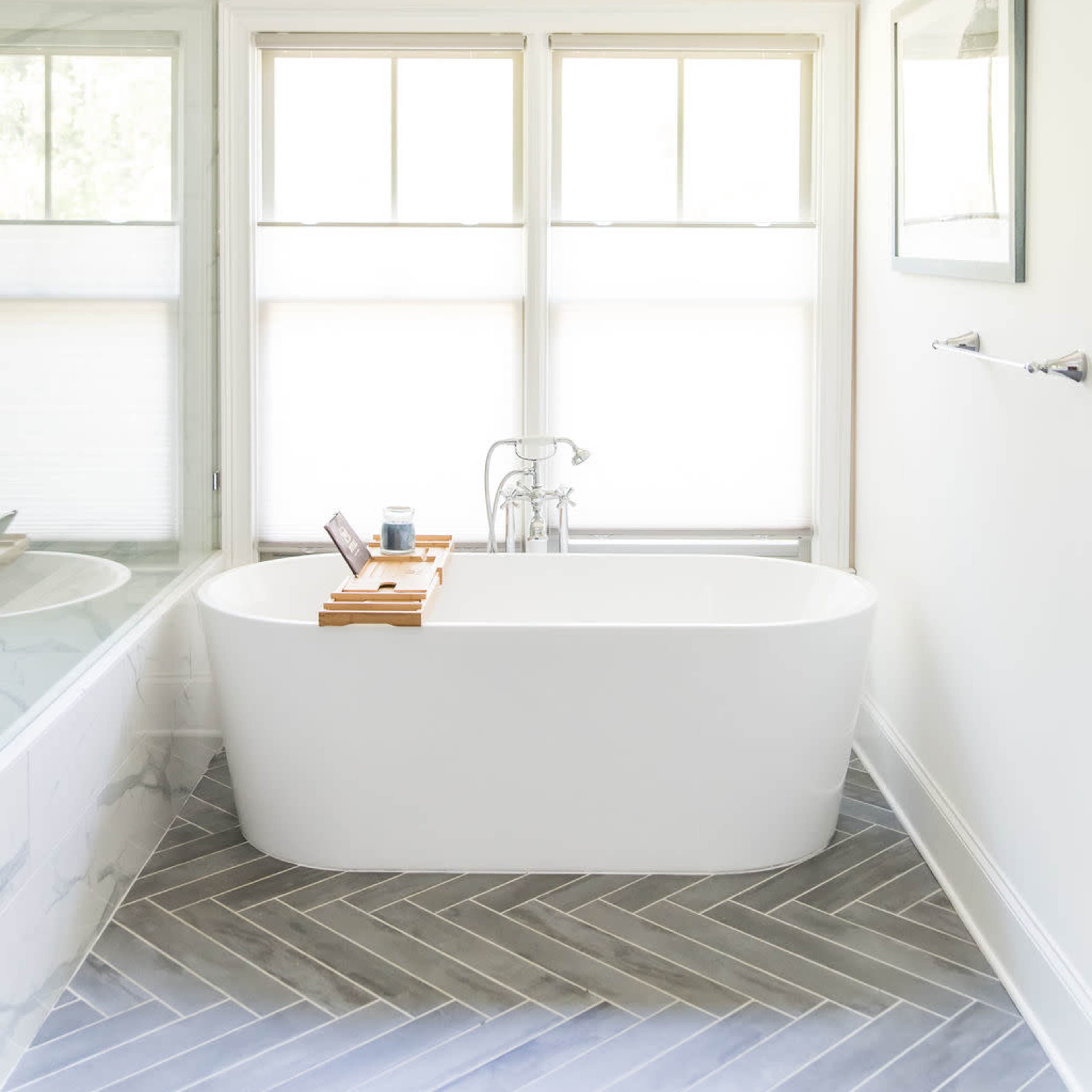 The bathroom features a freestanding white bathtub, a wooden bath tray, and large windows with blinds, all set against herringbone-patterned flooring.