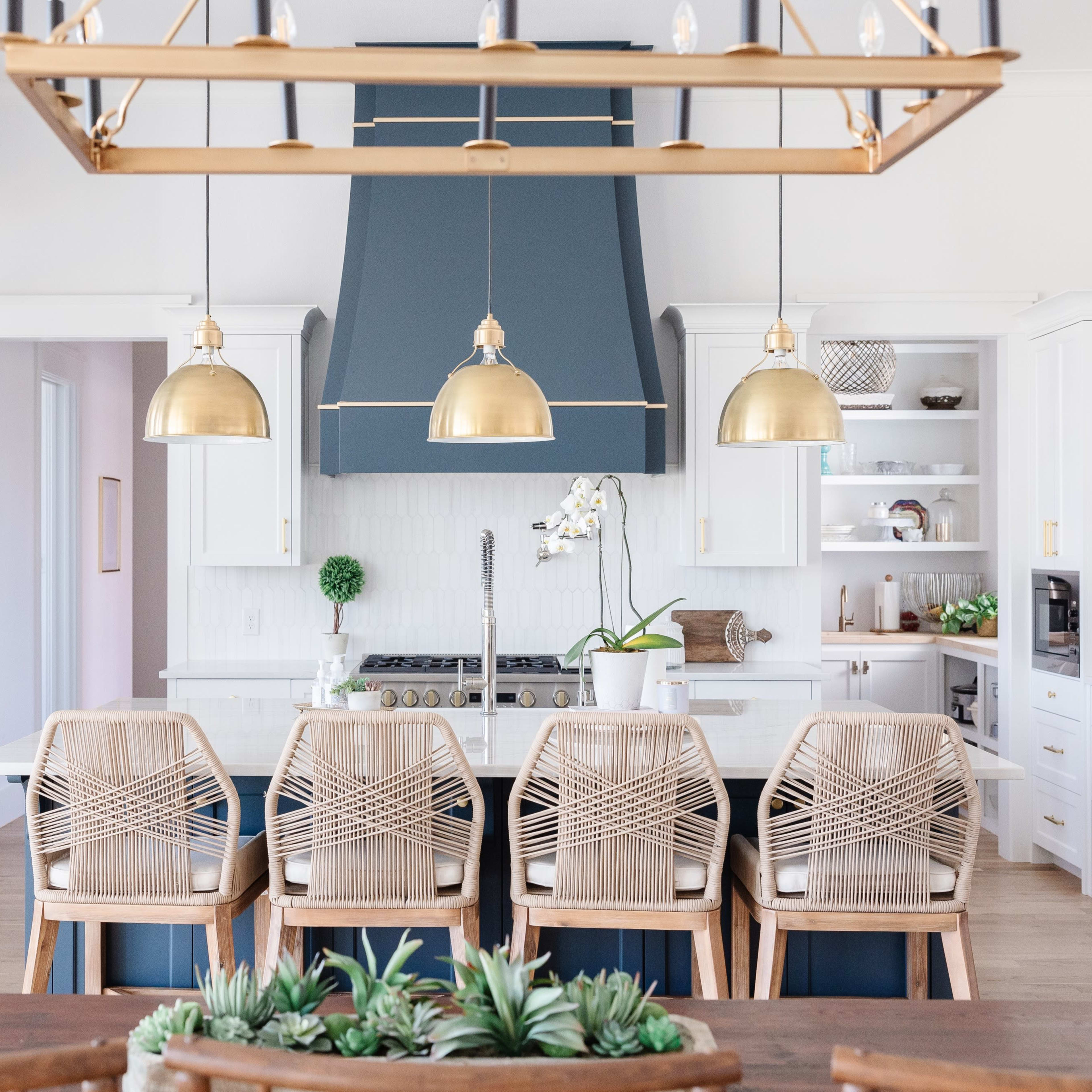 A modern kitchen with a large island, gold pendant lights, and wooden chairs arranged around a dining table.