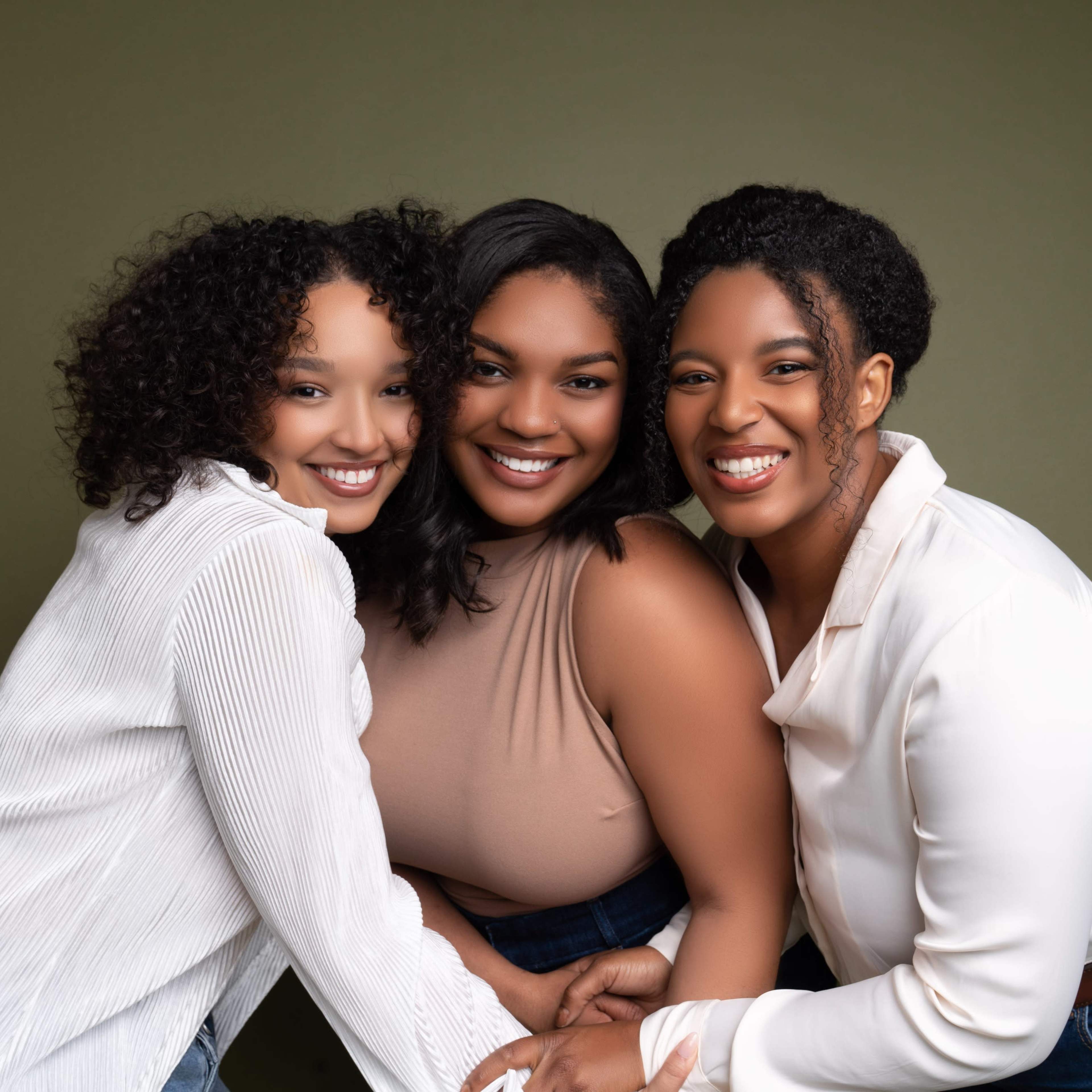 Three women stand closely together, smiling at the camera against a green background.