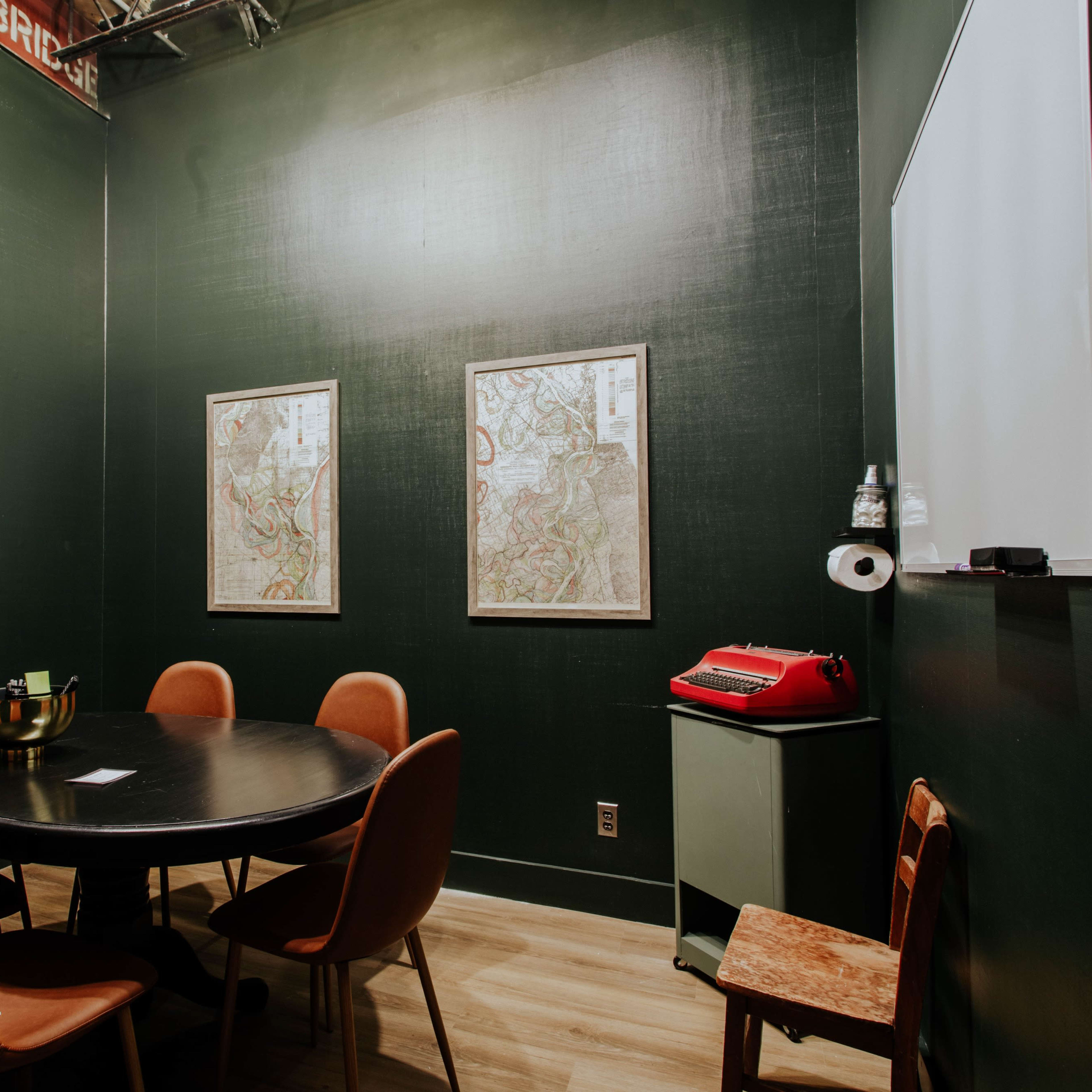 A small, dark green meeting room with a round table, four chairs, a television on the wall, and two framed maps.