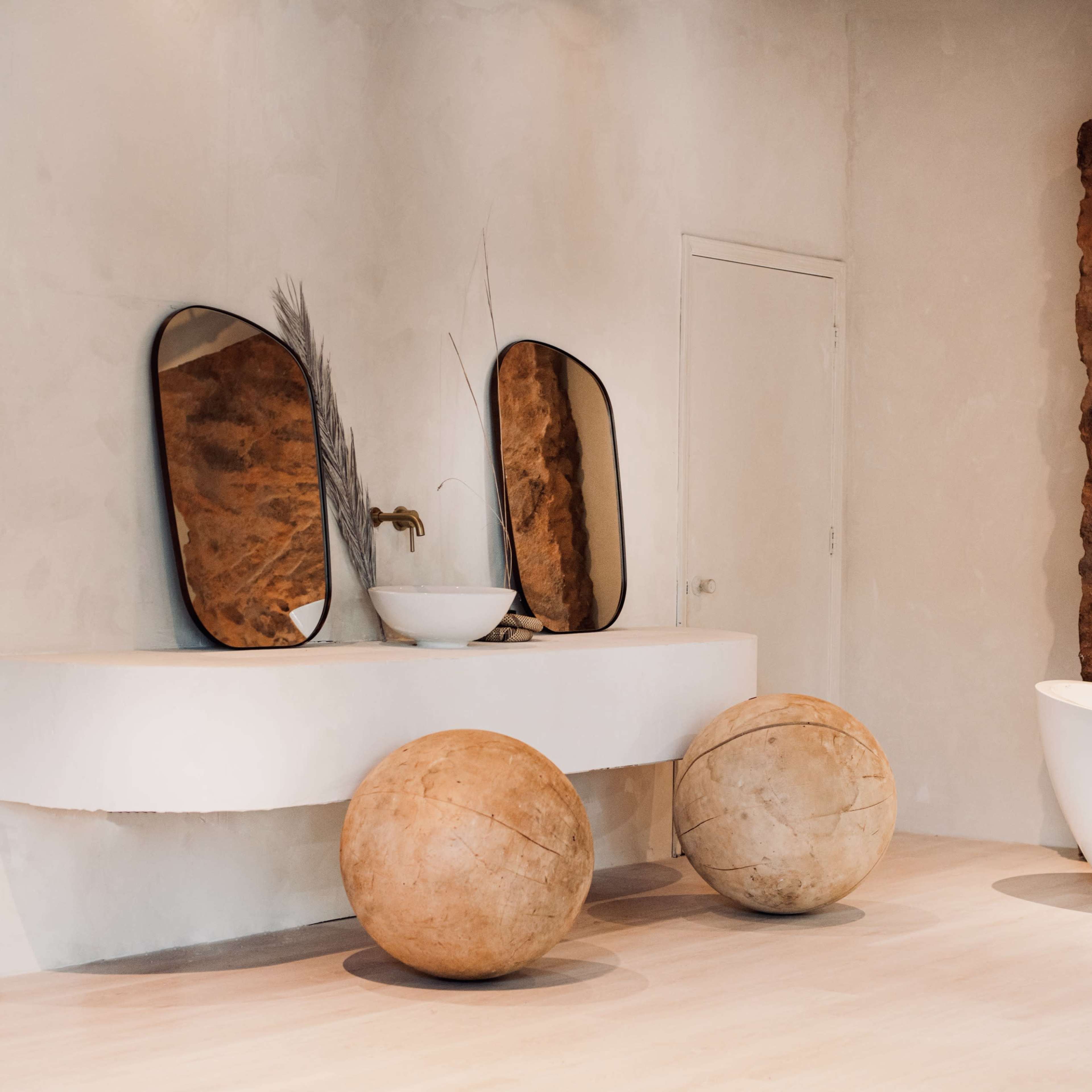 A minimalist bathroom with a stone wall, two circular wooden balls, a curved vanity with two mirrors, and a freestanding bathtub.