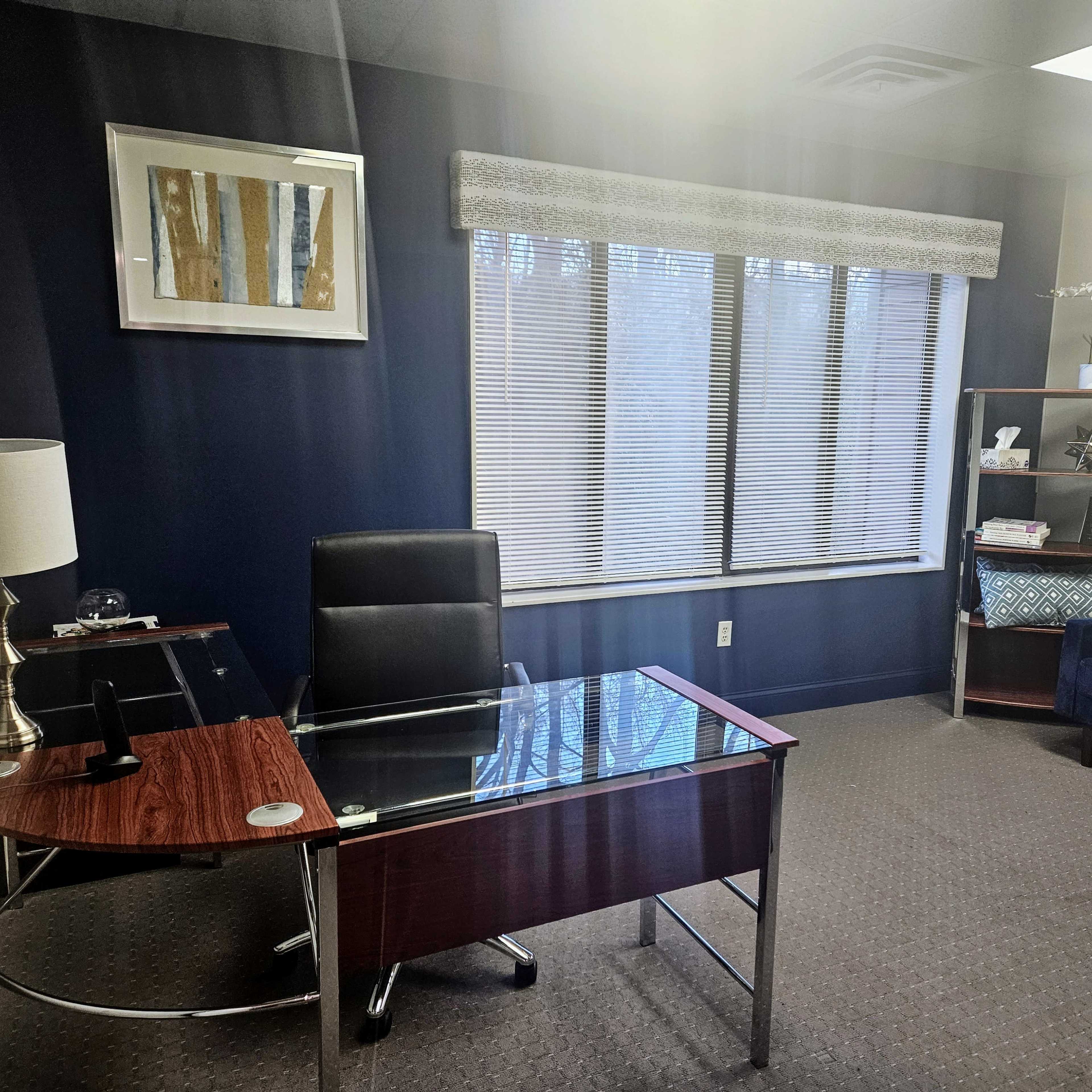 An office space featuring a glass-top desk, a black chair, a bookshelf, and a blue sofa, with large windows allowing light to enter.
