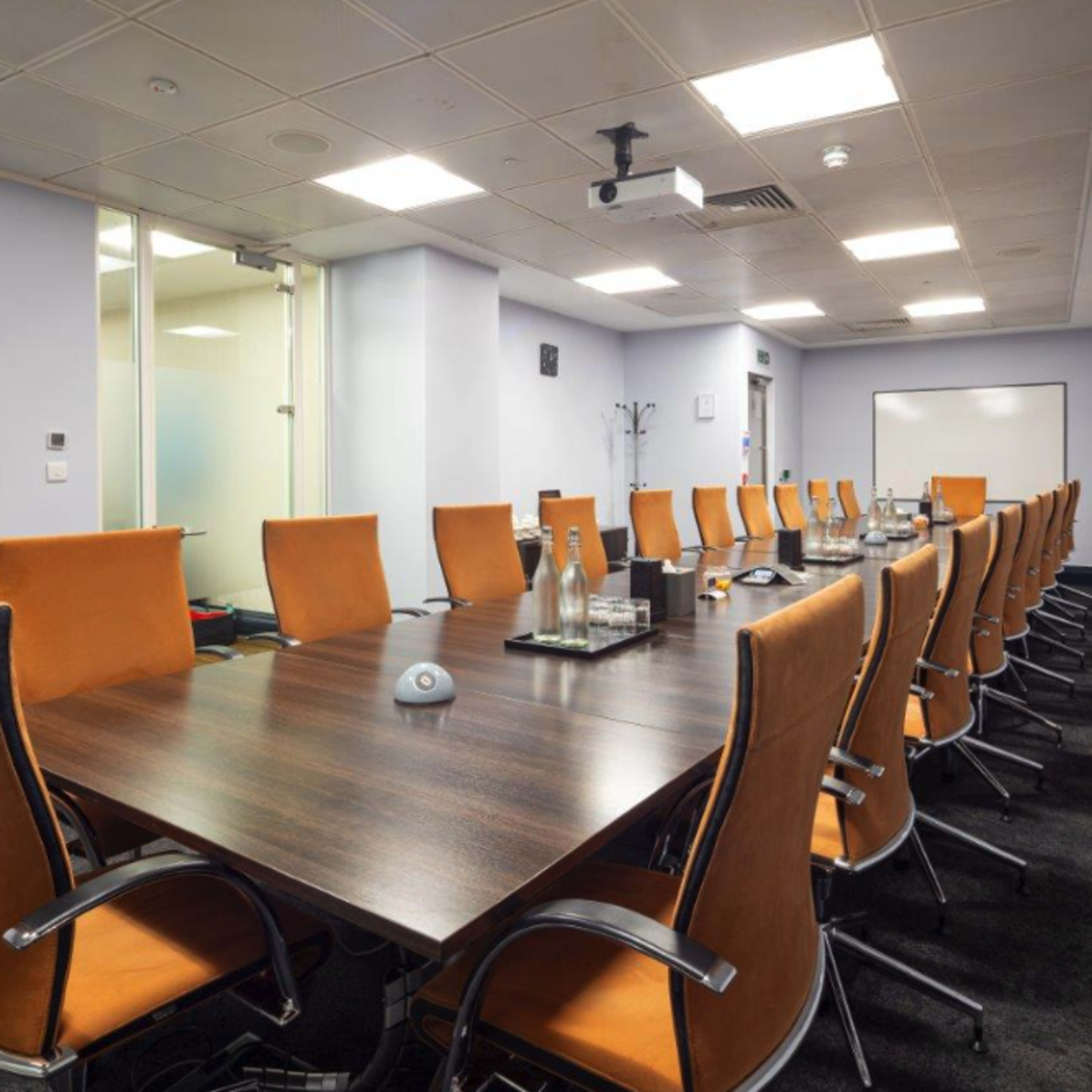 A large conference room features a long wooden table surrounded by orange chairs, with a projector and whiteboard visible in the background.