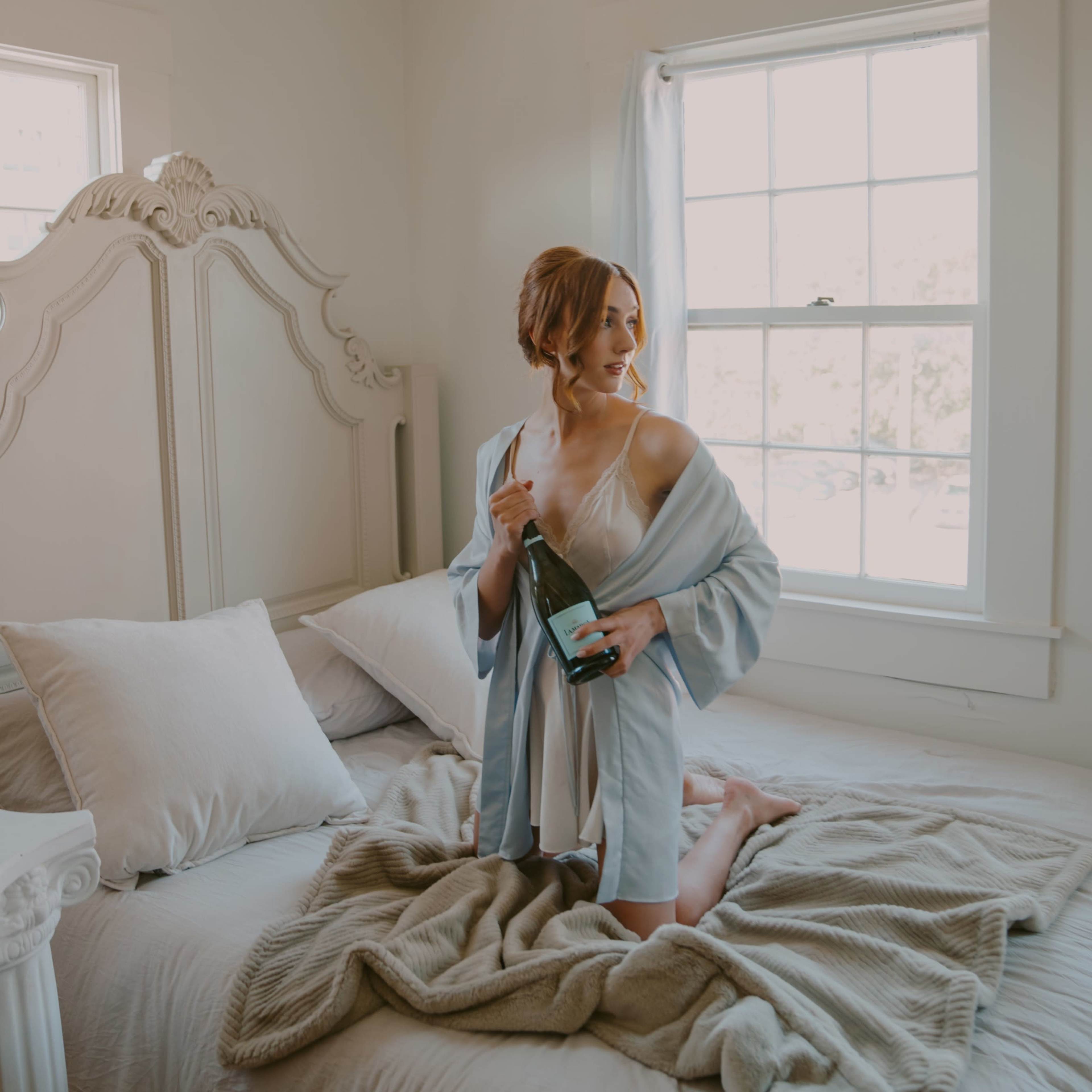 A woman stands on a bed in a well-lit room, holding a bottle of champagne while wrapped in a light-colored robe.