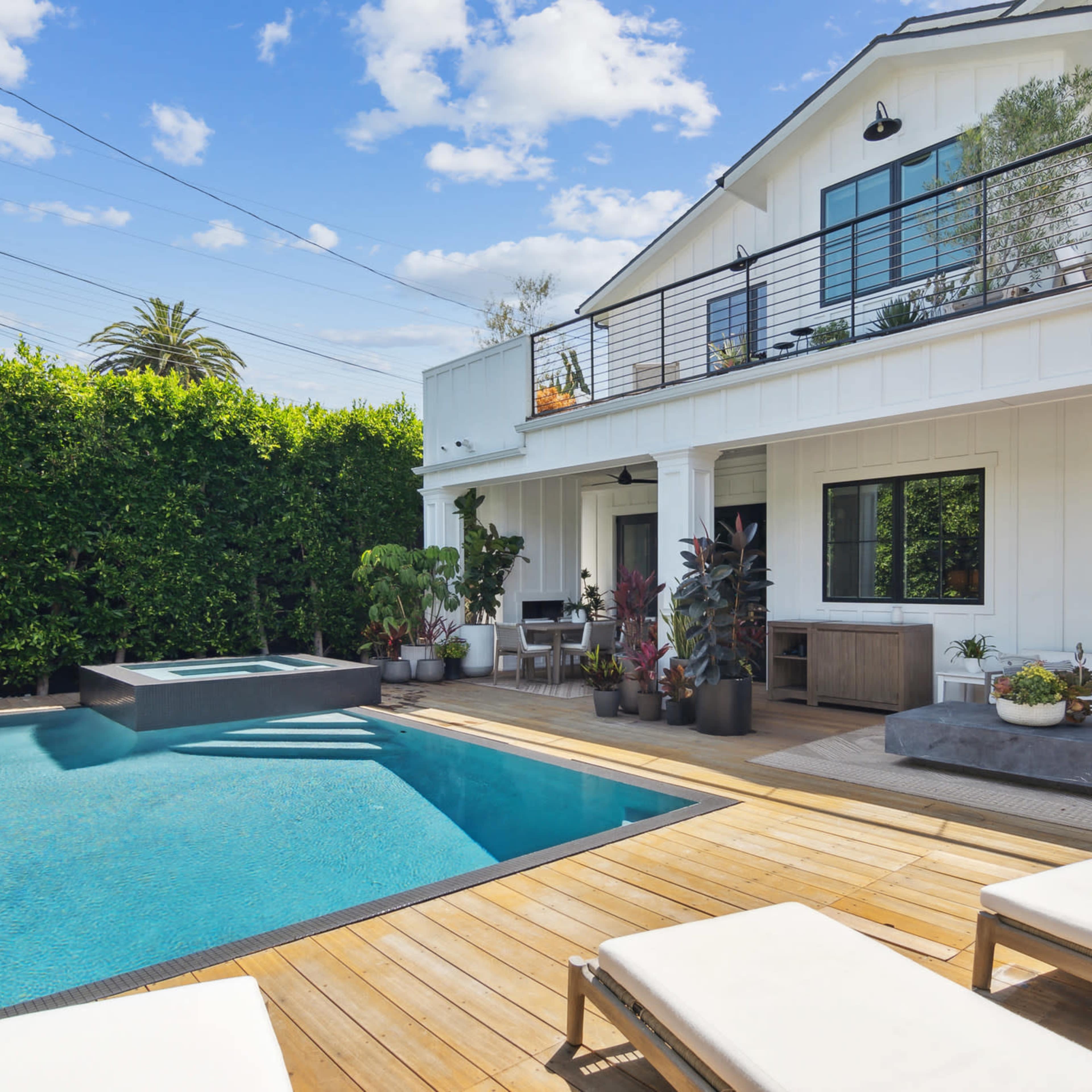 A modern outdoor area featuring a rectangular swimming pool surrounded by wooden decking, potted plants, and lounge chairs, alongside a white house with large windows and a second-floor balcony.