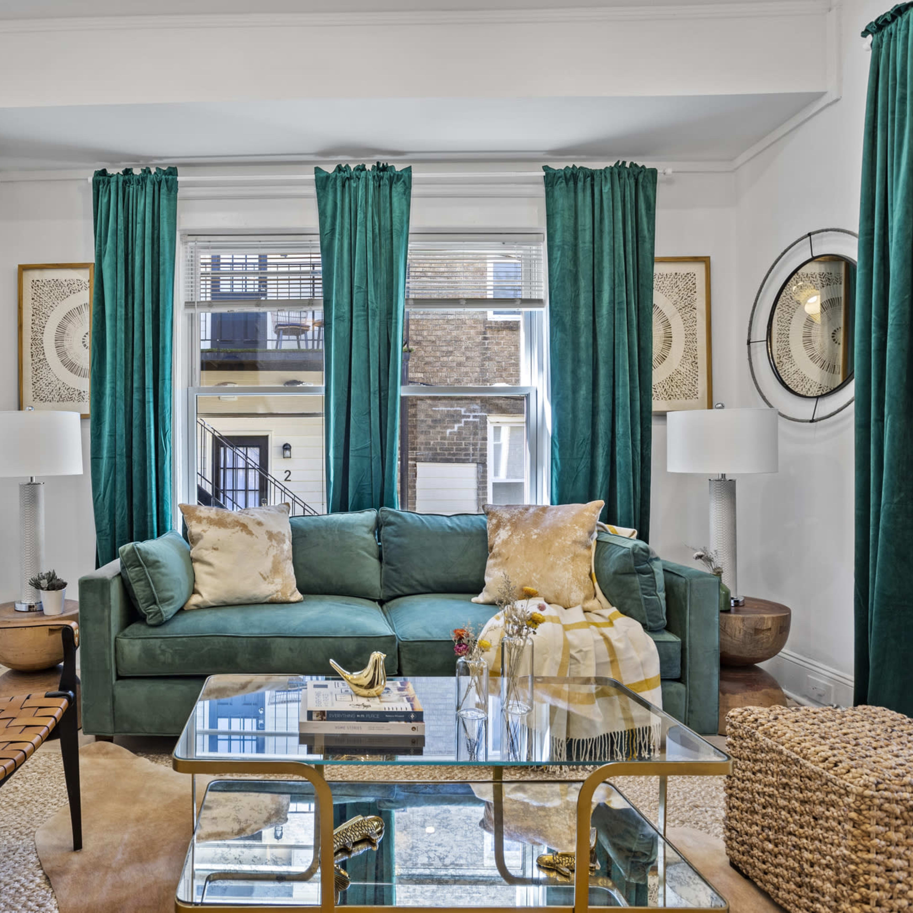 A living room featuring a green sofa, a glass coffee table, and large windows framed by teal curtains.