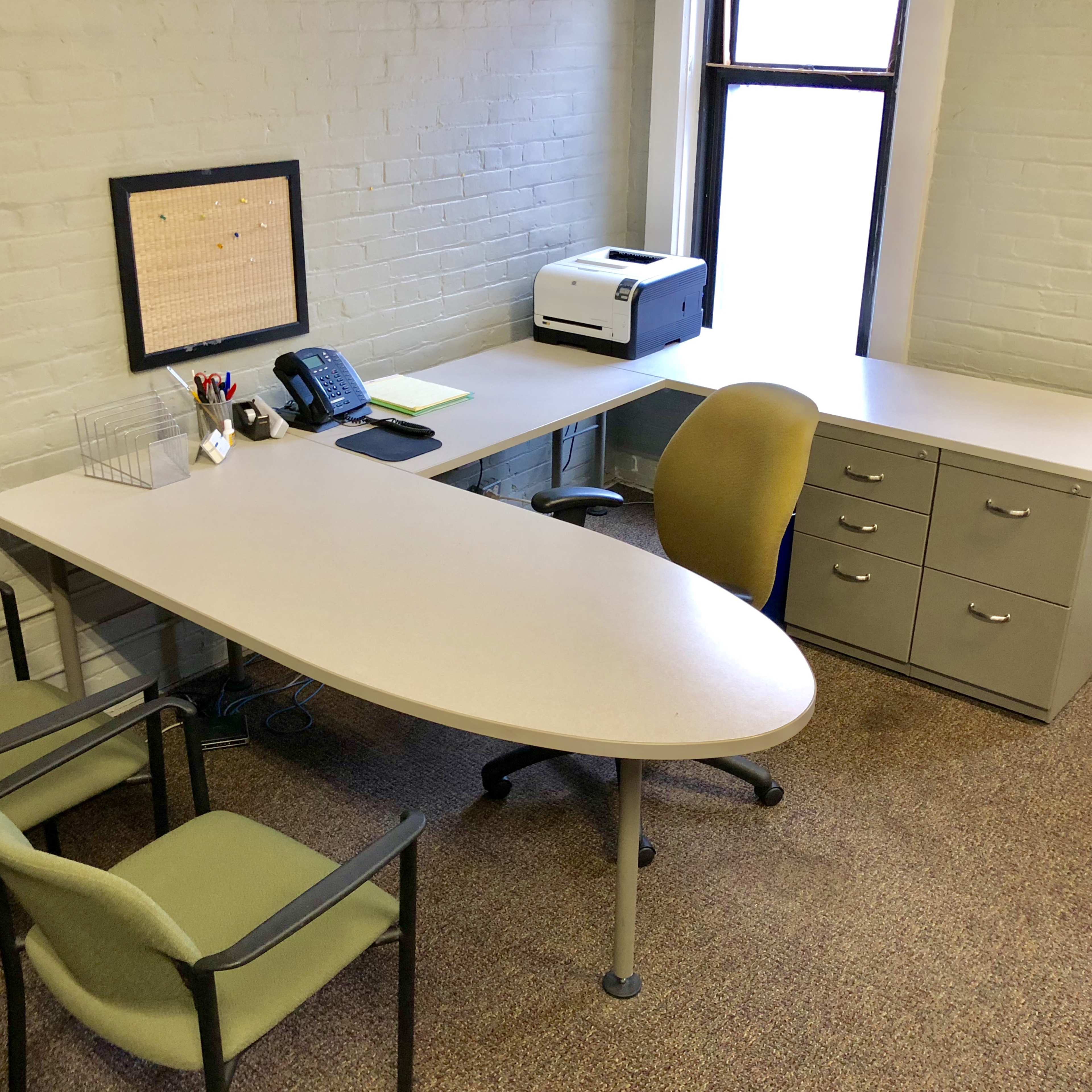 A small office space with a curved desk, a printer, a phone, two green chairs, and a filing cabinet against a light-colored brick wall.