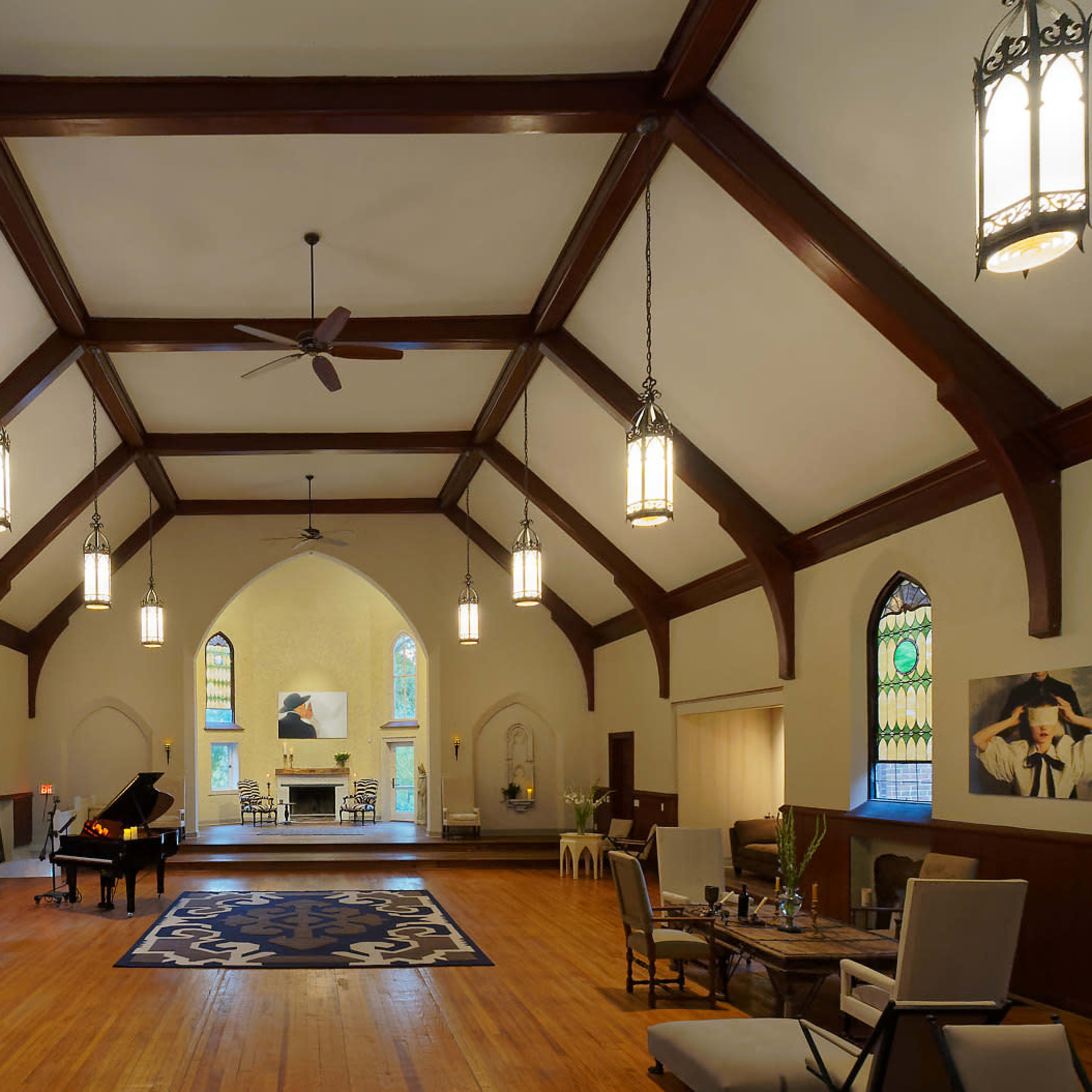 The interior of a spacious hall features wooden beams, large windows with stained glass, and a black piano positioned on a light-colored floor.