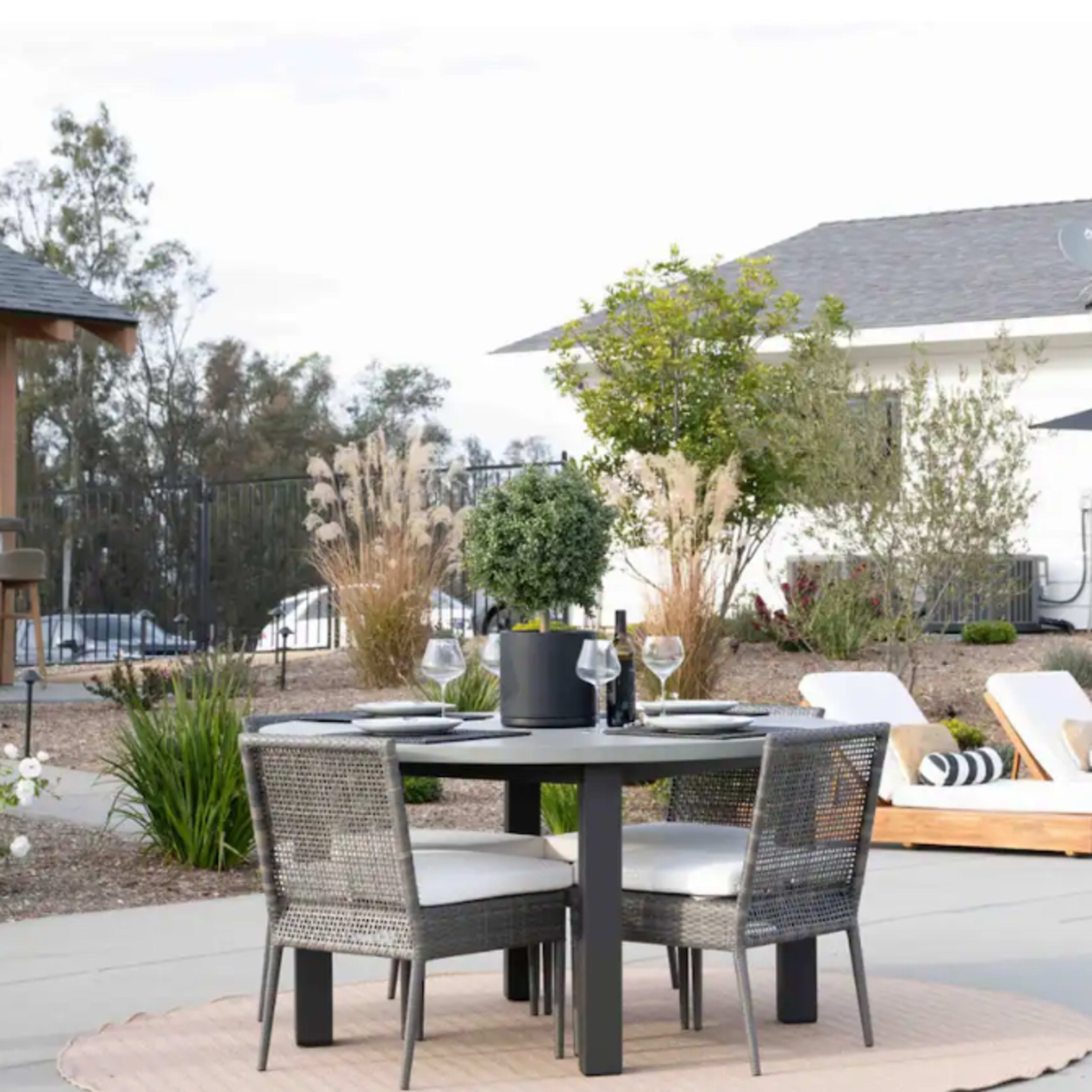 A patio features a round dining table with four chairs, a large umbrella, and two lounge chairs beside a small pool, set against a modern white house.
