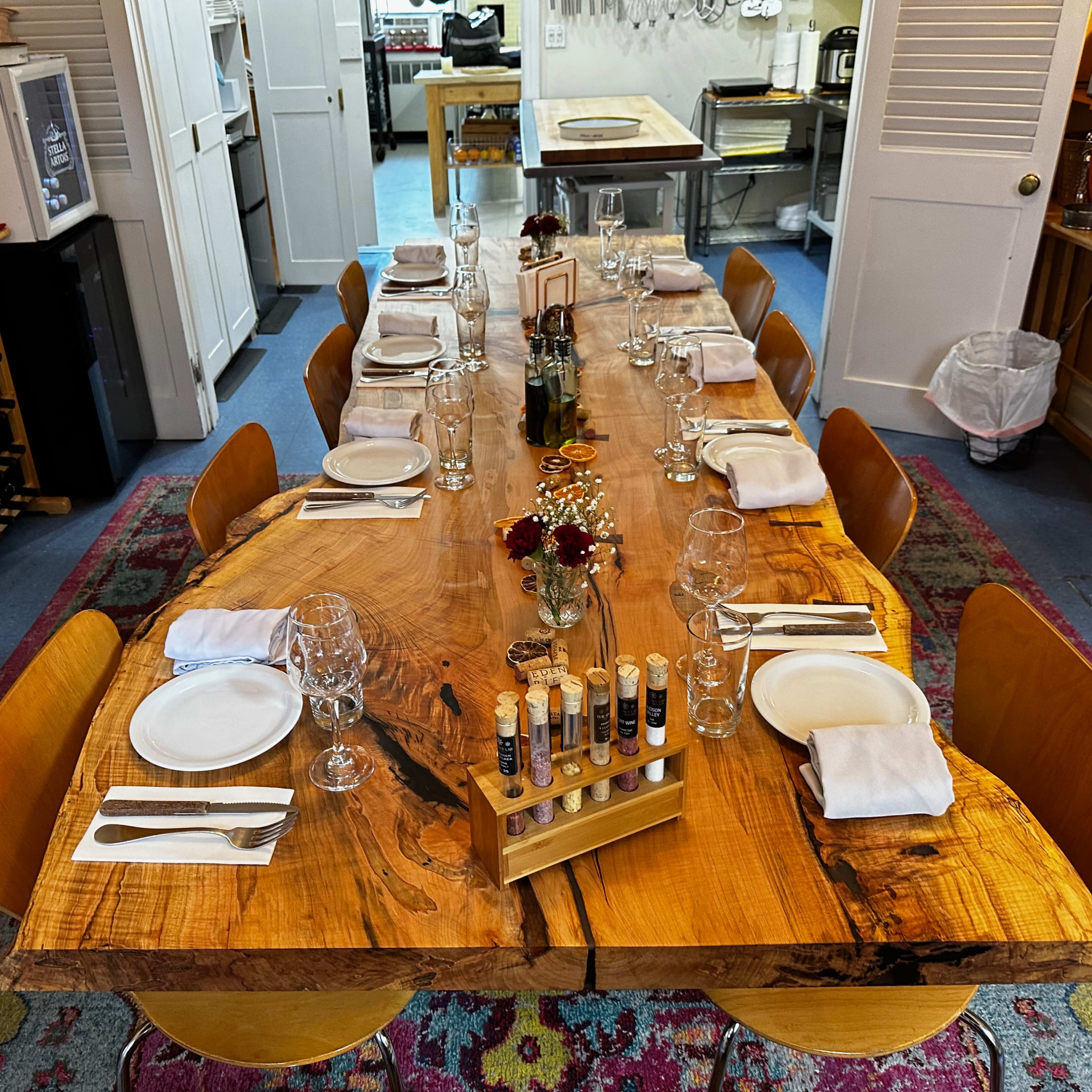 A long wooden dining table is set with plates, glasses, and utensils, surrounded by chairs in a kitchen environment.