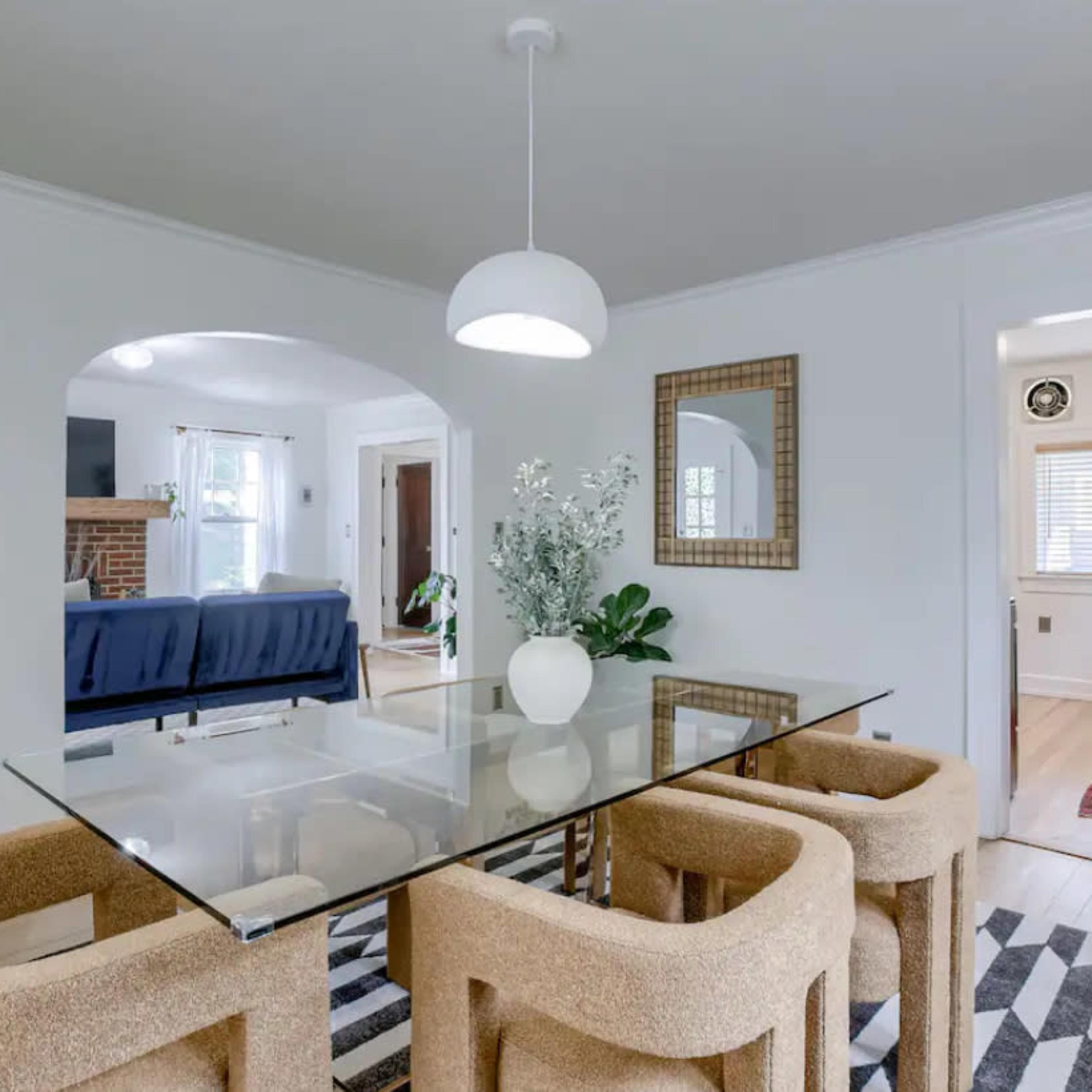A dining area features a glass table surrounded by upholstered chairs, with a decorative plant and mirror in the backdrop.