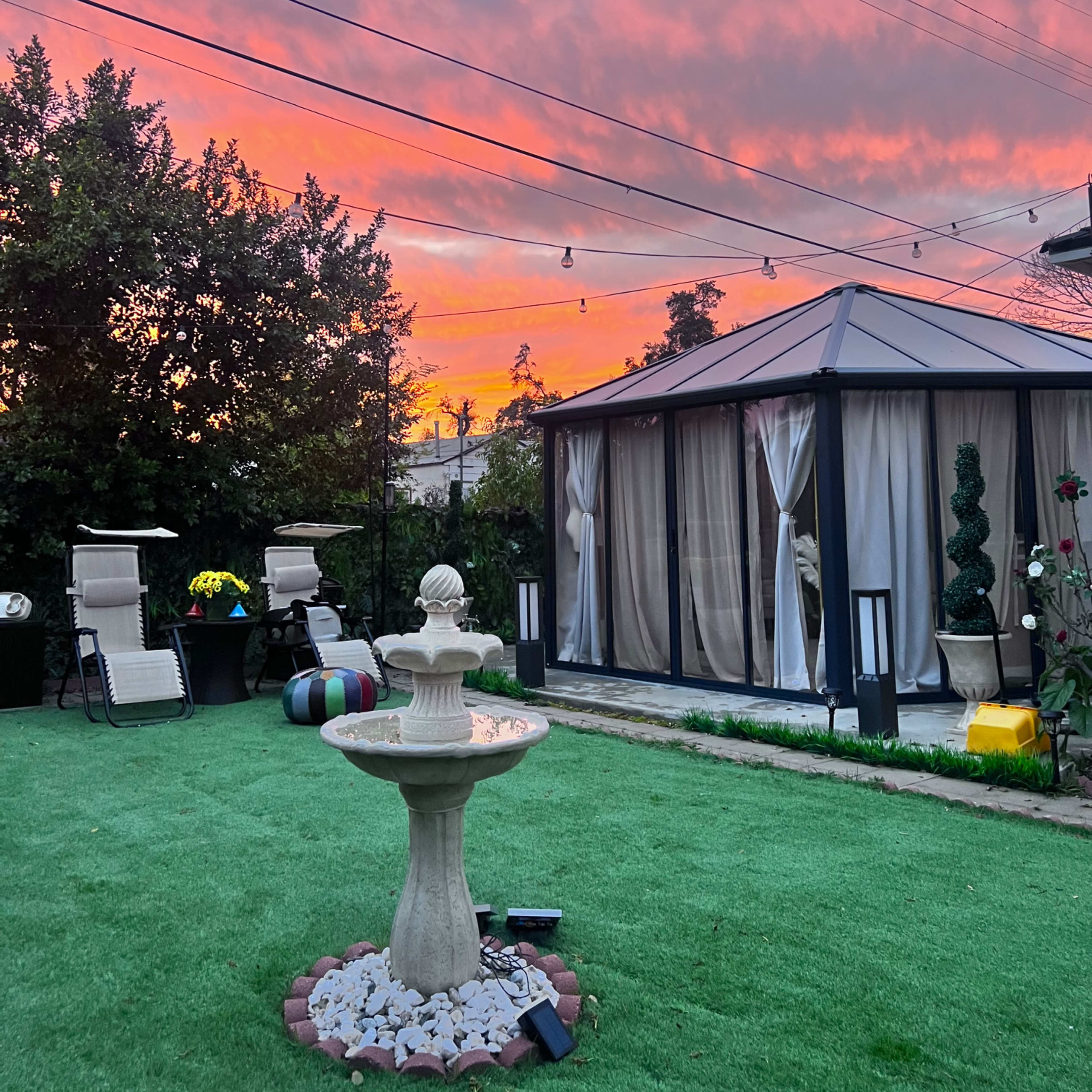 The scene features a landscaped backyard with a fountain in the center, surrounded by chairs and a gazebo, under a colorful sunset sky.