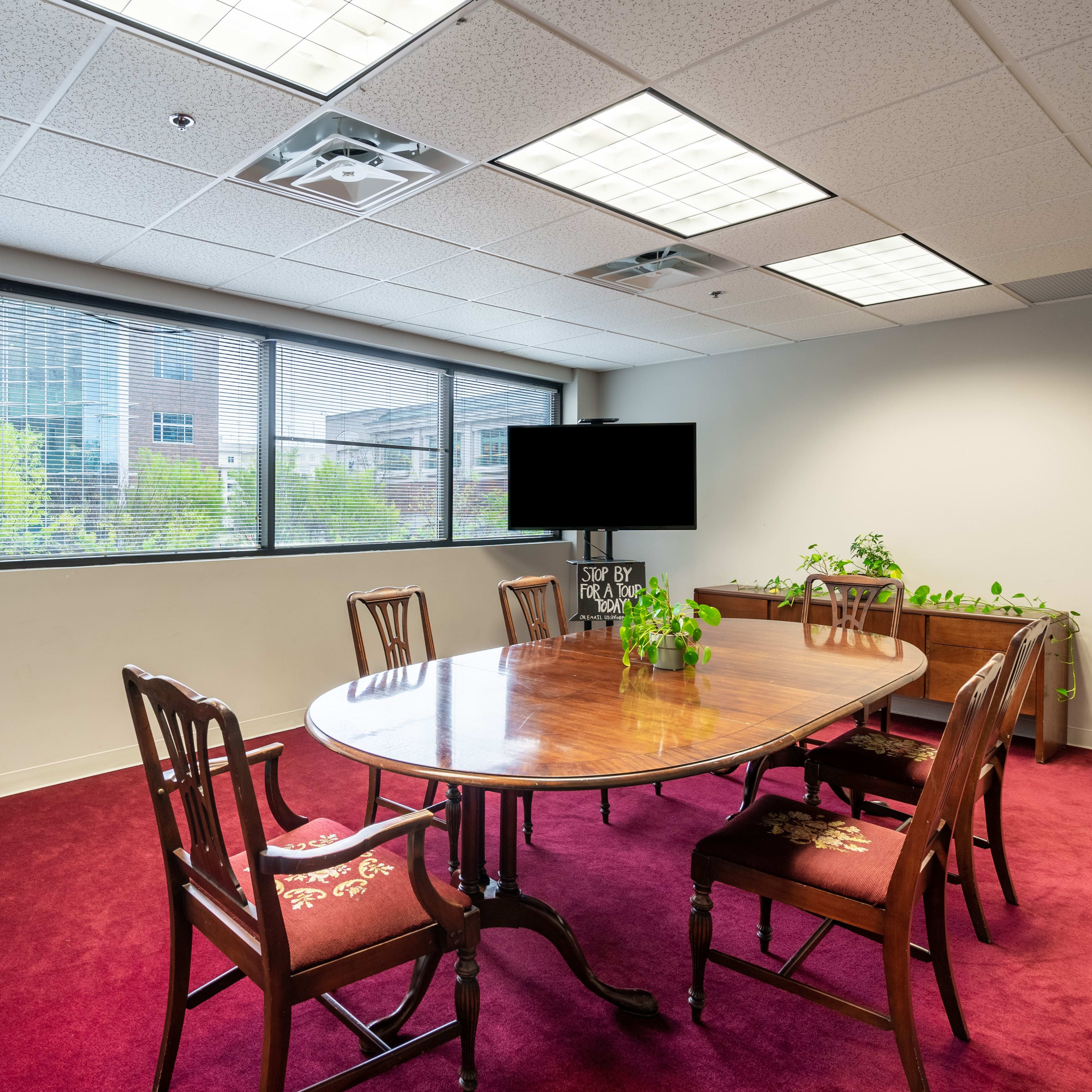A conference room features a long wooden table surrounded by chairs, with large windows providing natural light and plants placed around the space.