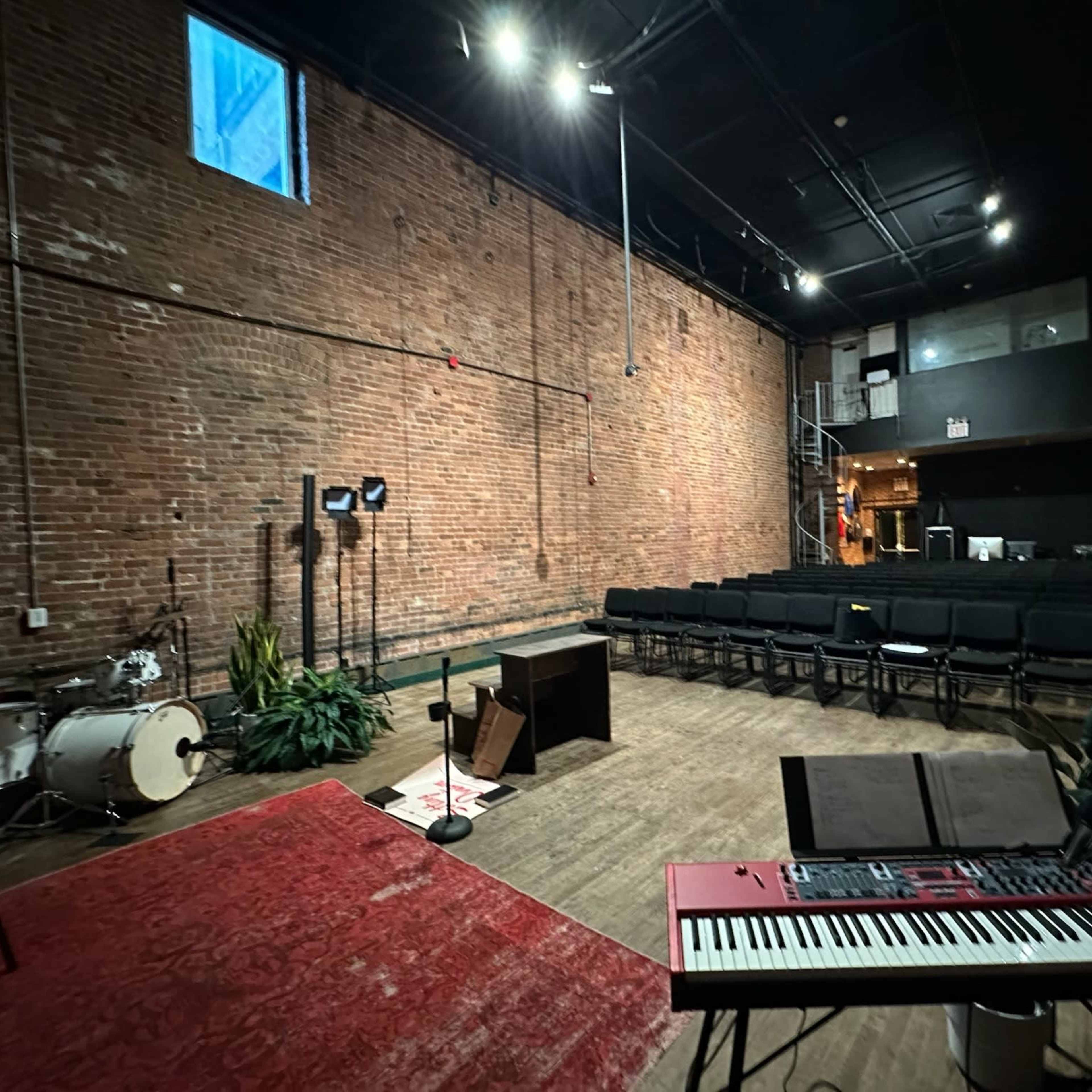 An empty music venue featuring a brick wall, a keyboard, a microphone stand, and rows of black chairs arranged for an audience.