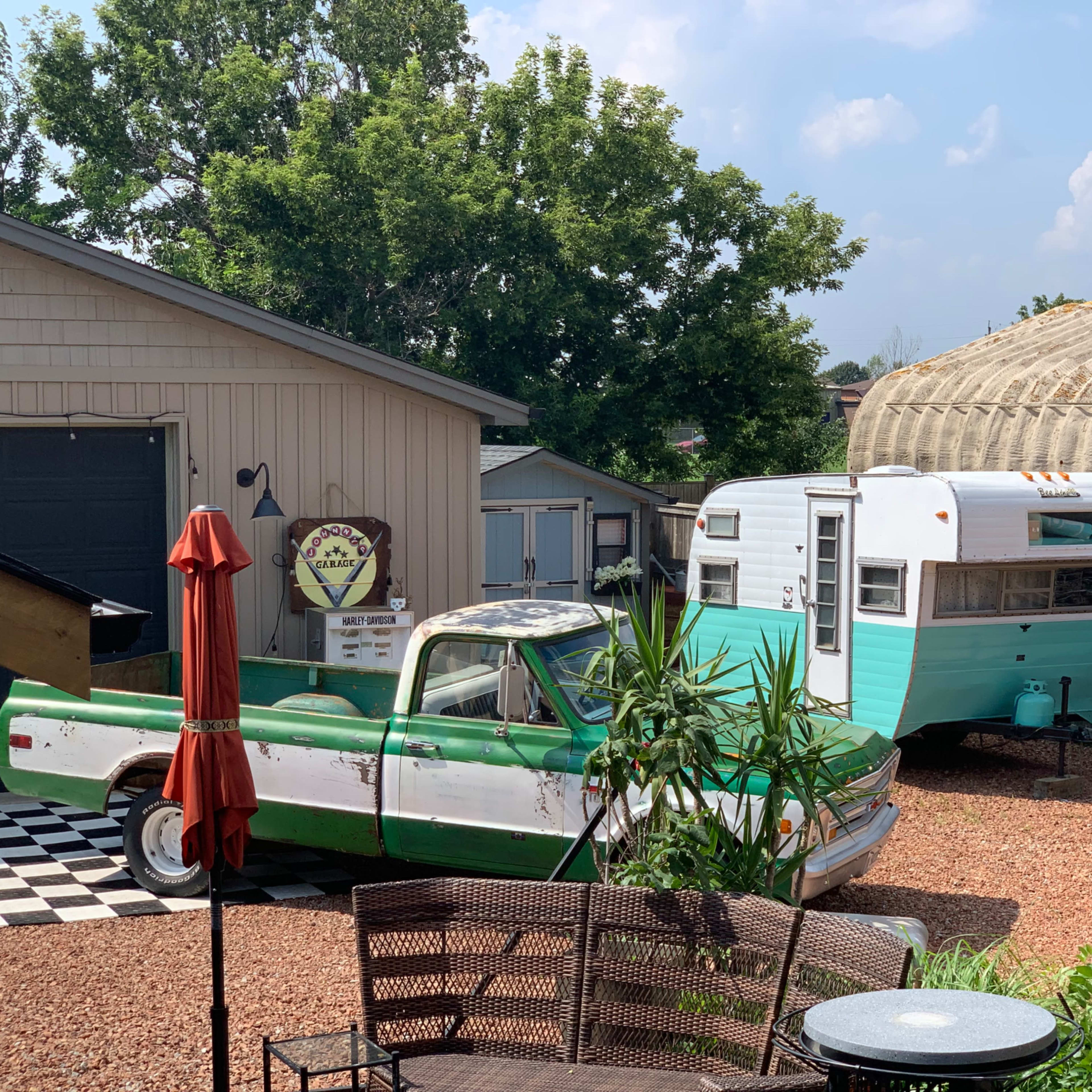 The scene features a vintage pickup truck parked beside a turquoise and white camper in a gravel yard with a checkered patio.