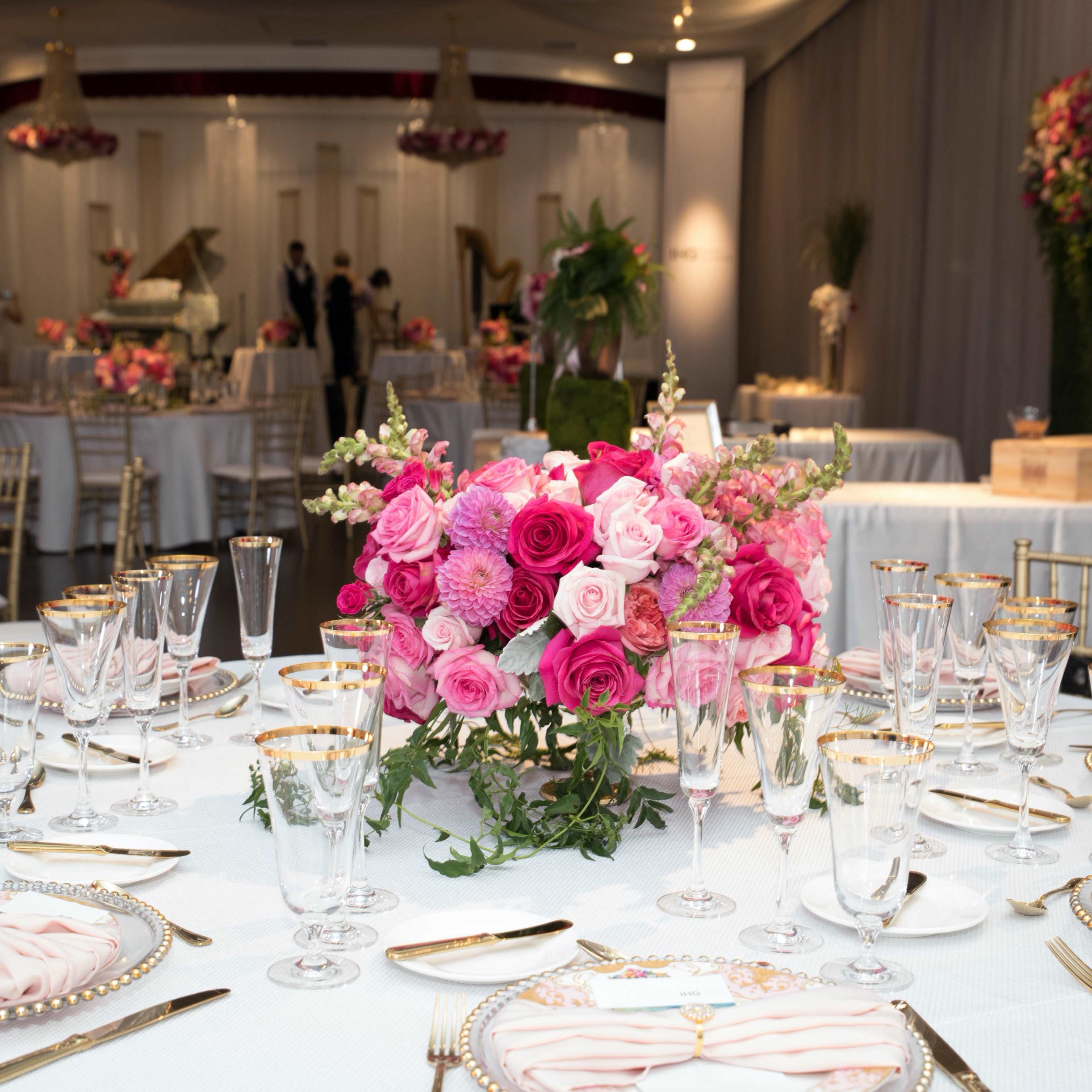 A elegantly set dining area features round tables adorned with floral centerpieces, fine china, and crystal glassware, with a backdrop of draped fabric and floral arrangements.