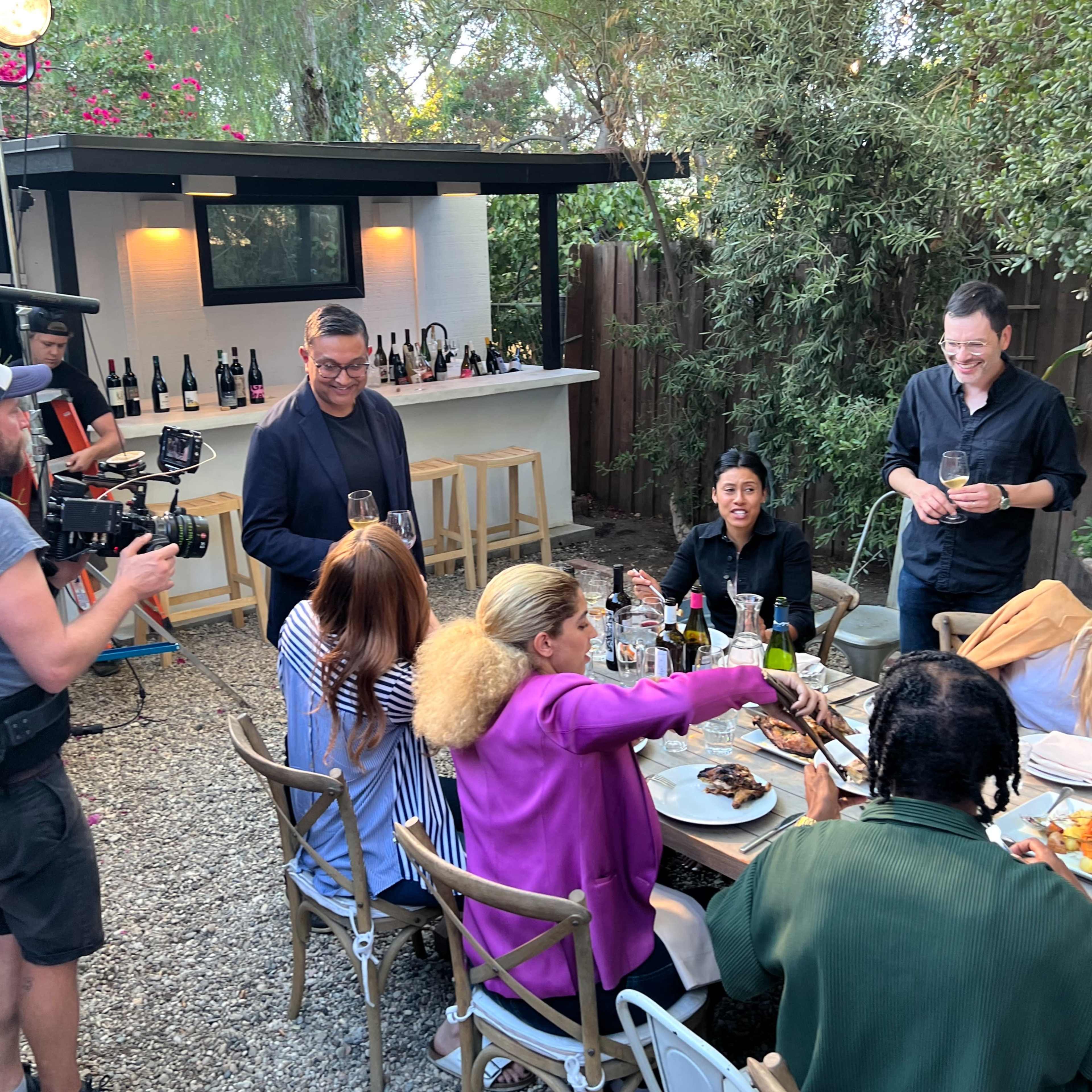 A group of people is gathered around a table in an outdoor setting, sharing food and drinks while a camera crew films the scene.