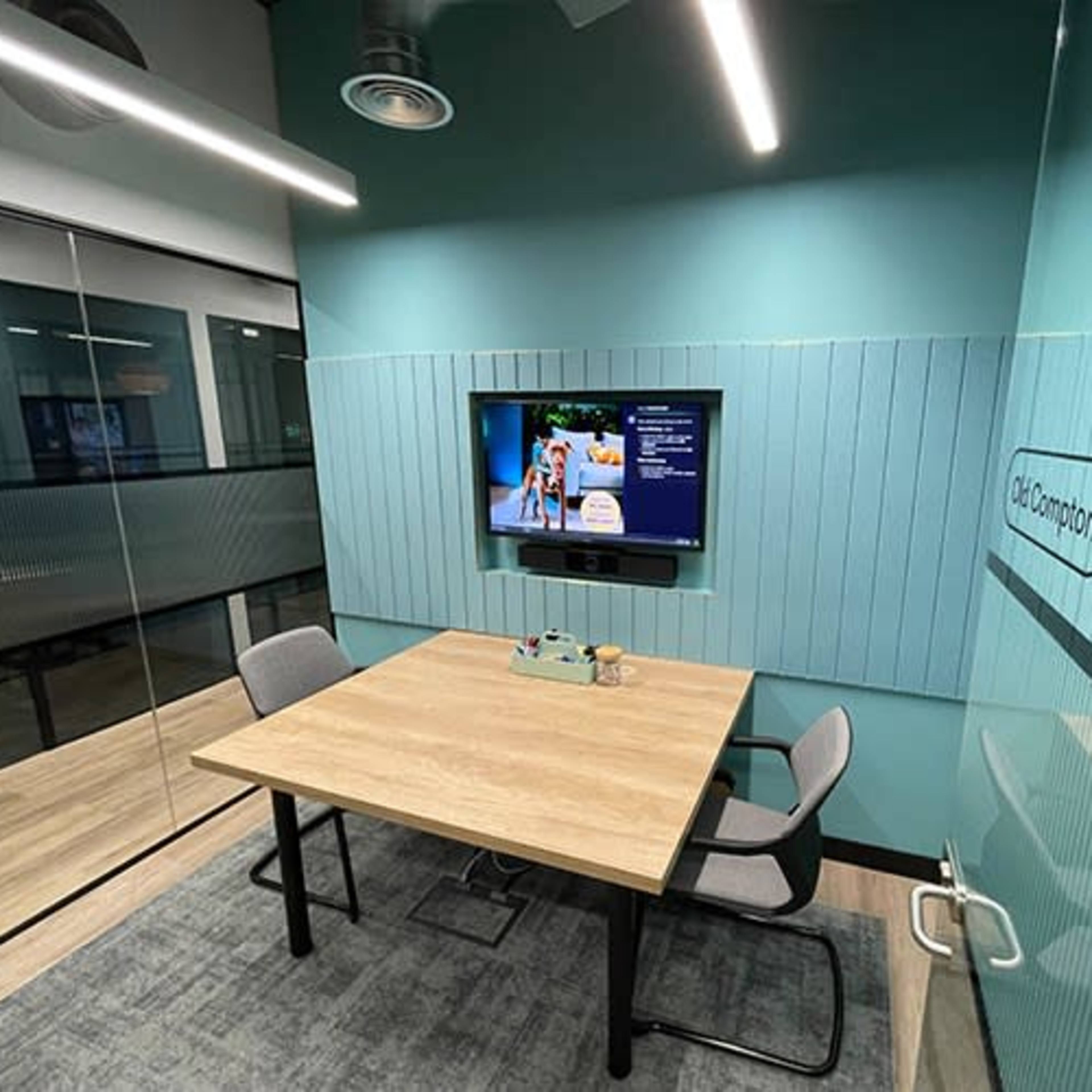 An office meeting room features a wooden table with two chairs, a television mounted on a wall, and glass partitions.
