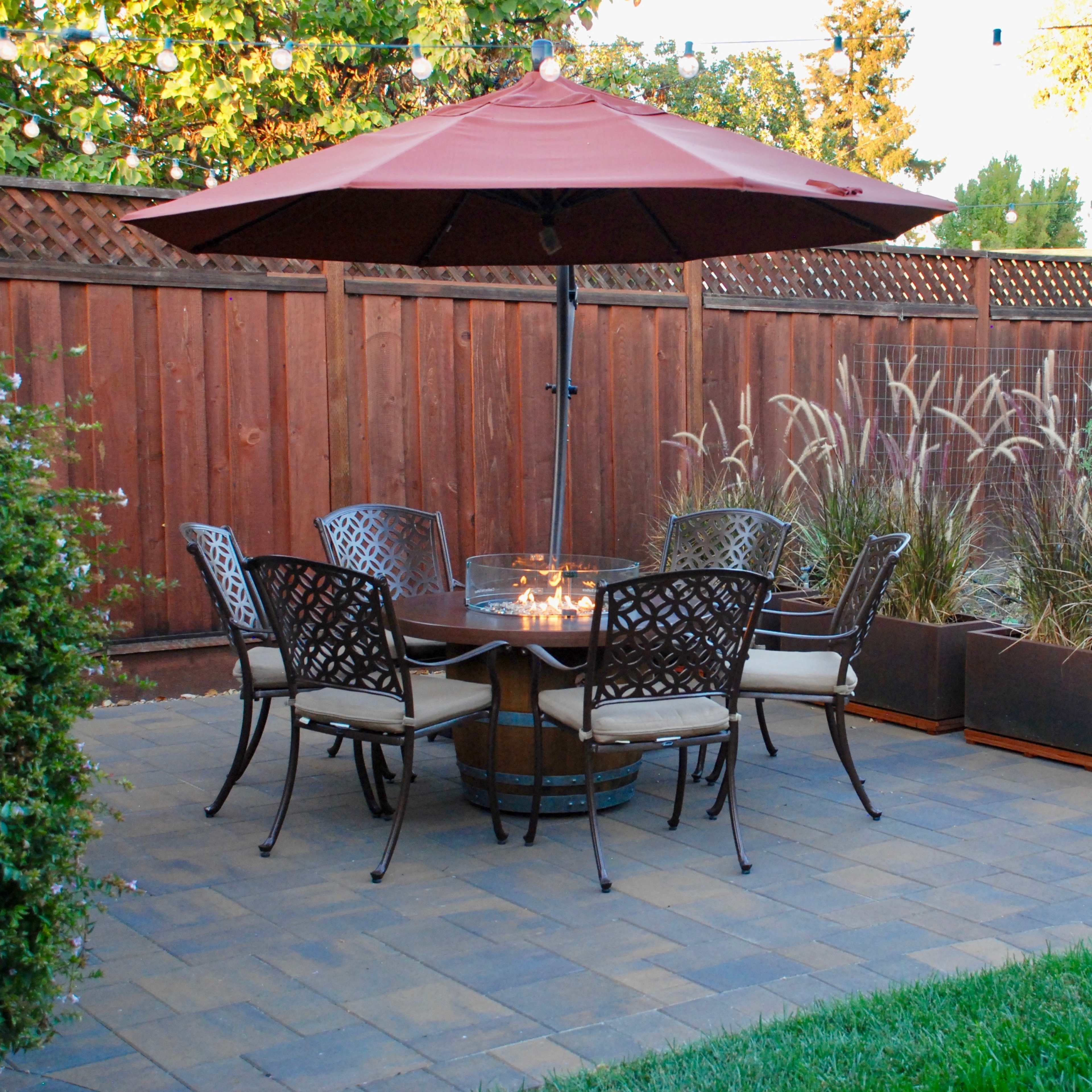 A patio set with six chairs and a central table, shaded by a large umbrella, is arranged on a paved surface surrounded by plants and a wooden fence.