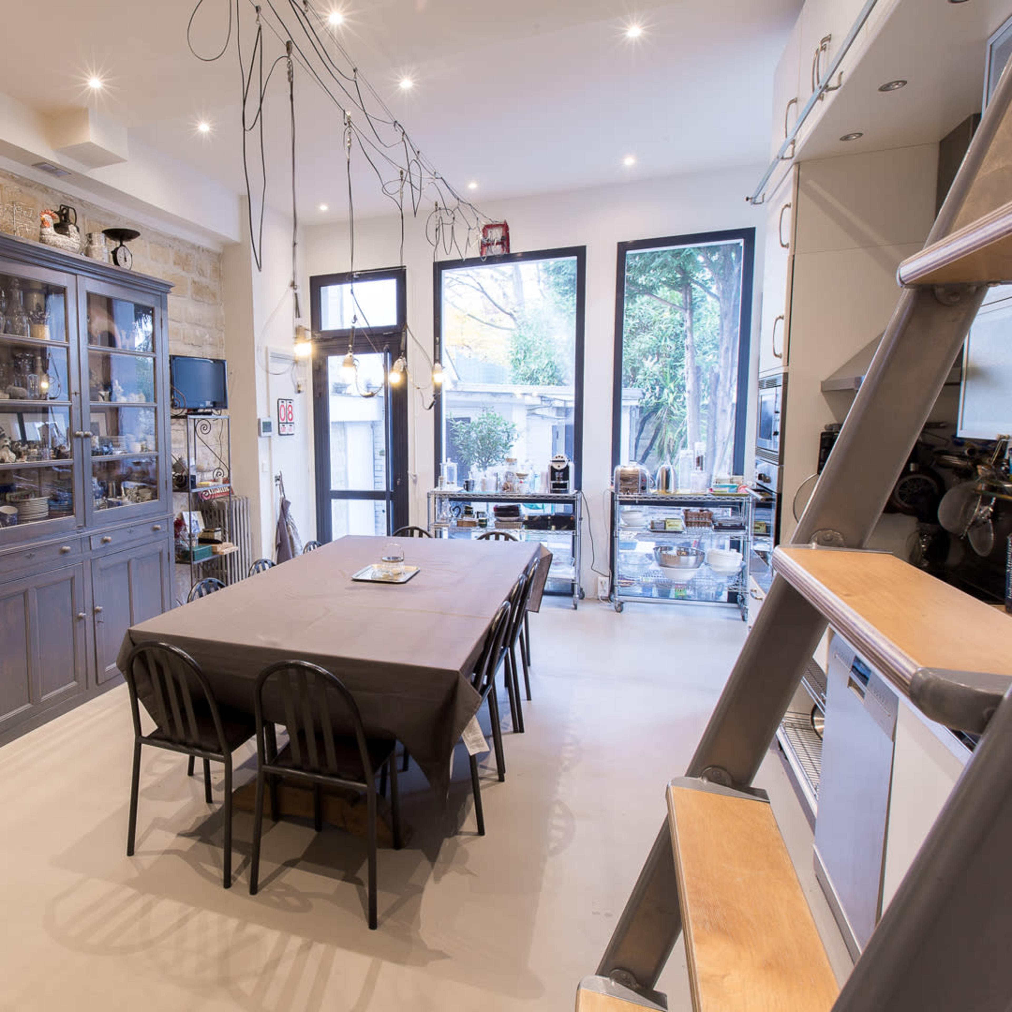A modern dining area with a long table, surrounded by chairs, next to a gray cabinet, and large windows leading to an outdoor space.