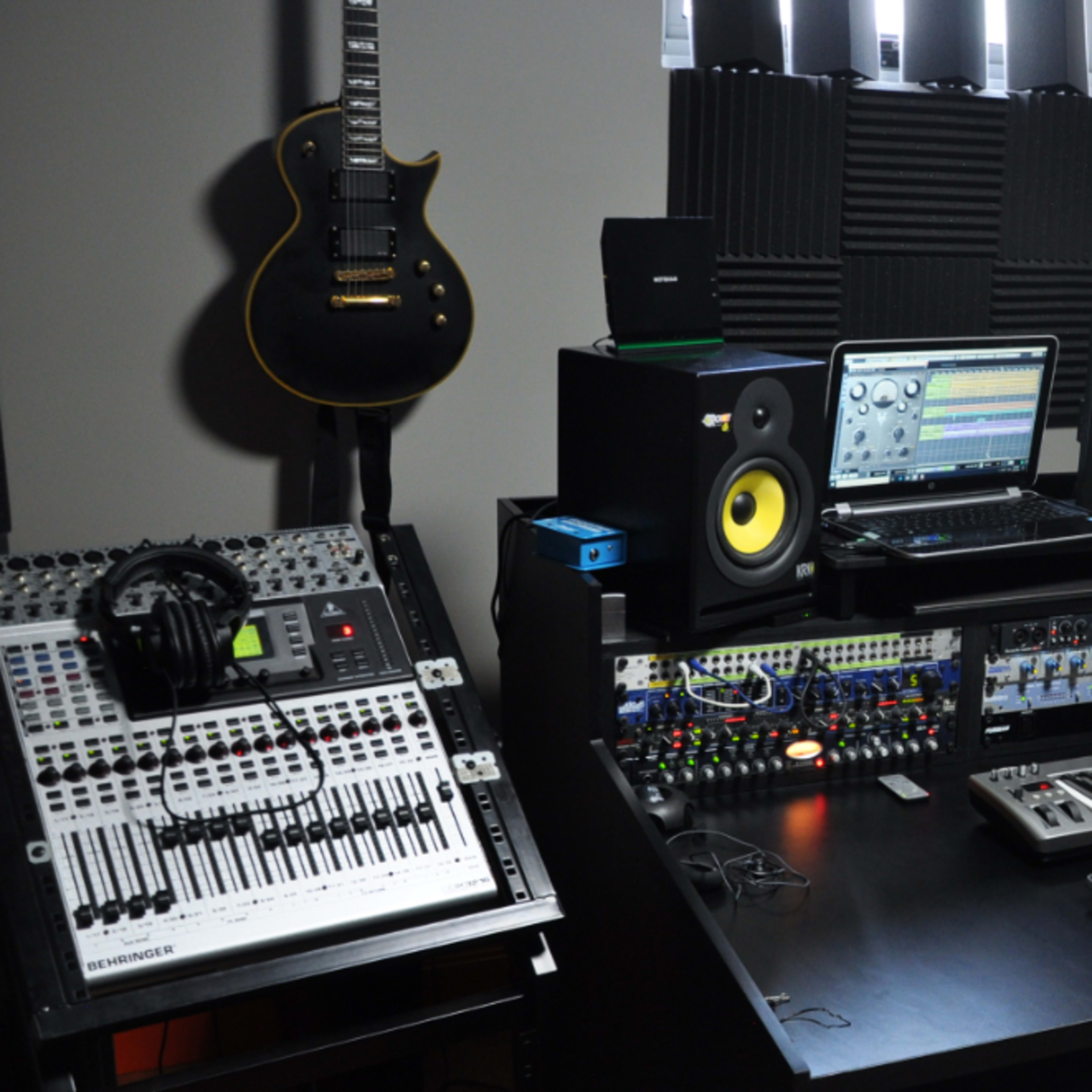 A music production studio featuring a desk with a computer, audio interface, synthesizers, speakers, a MIDI keyboard, and various music equipment, along with guitars mounted on the walls.