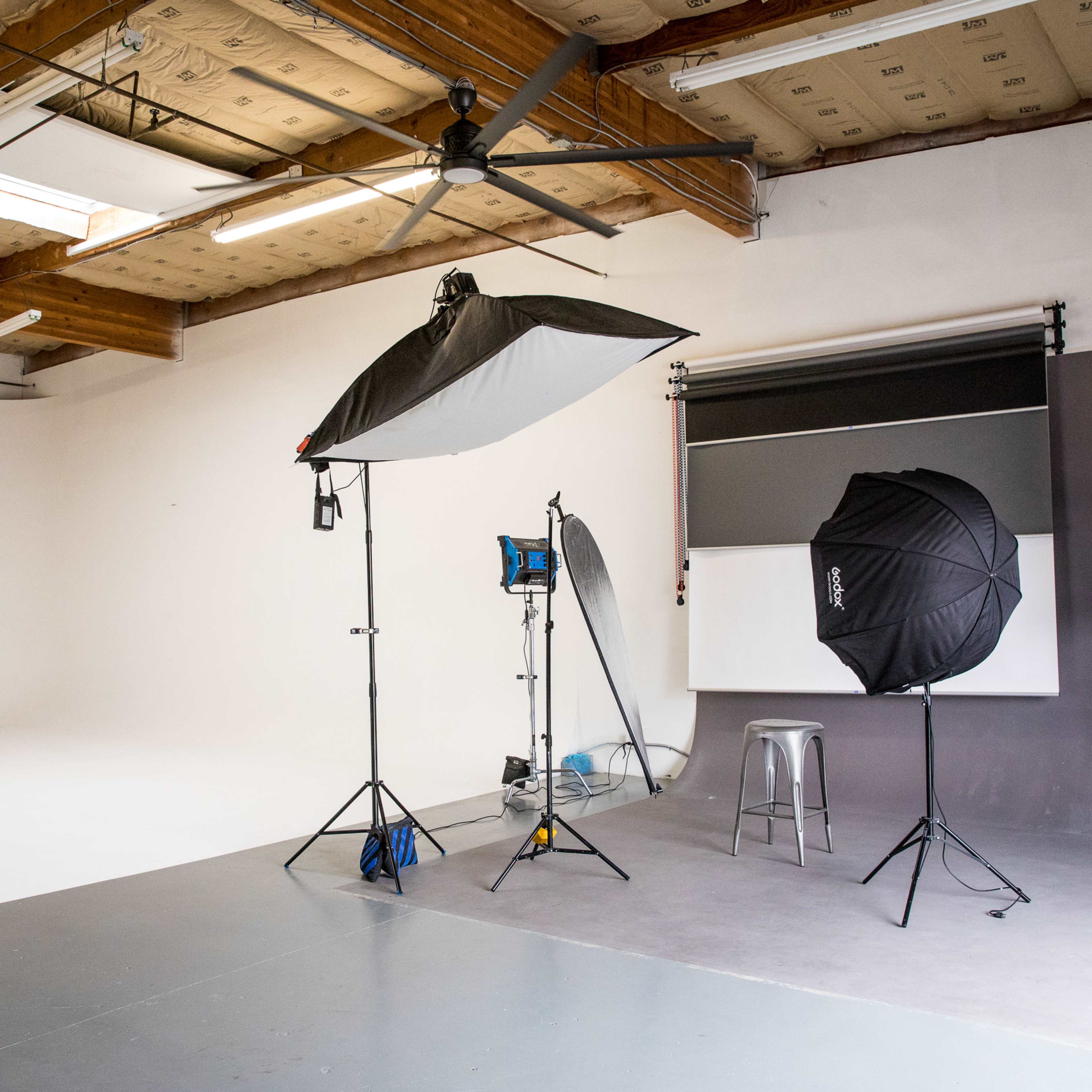 A photography studio features a white backdrop, lighting equipment on stands, and a stool against a plain wall.