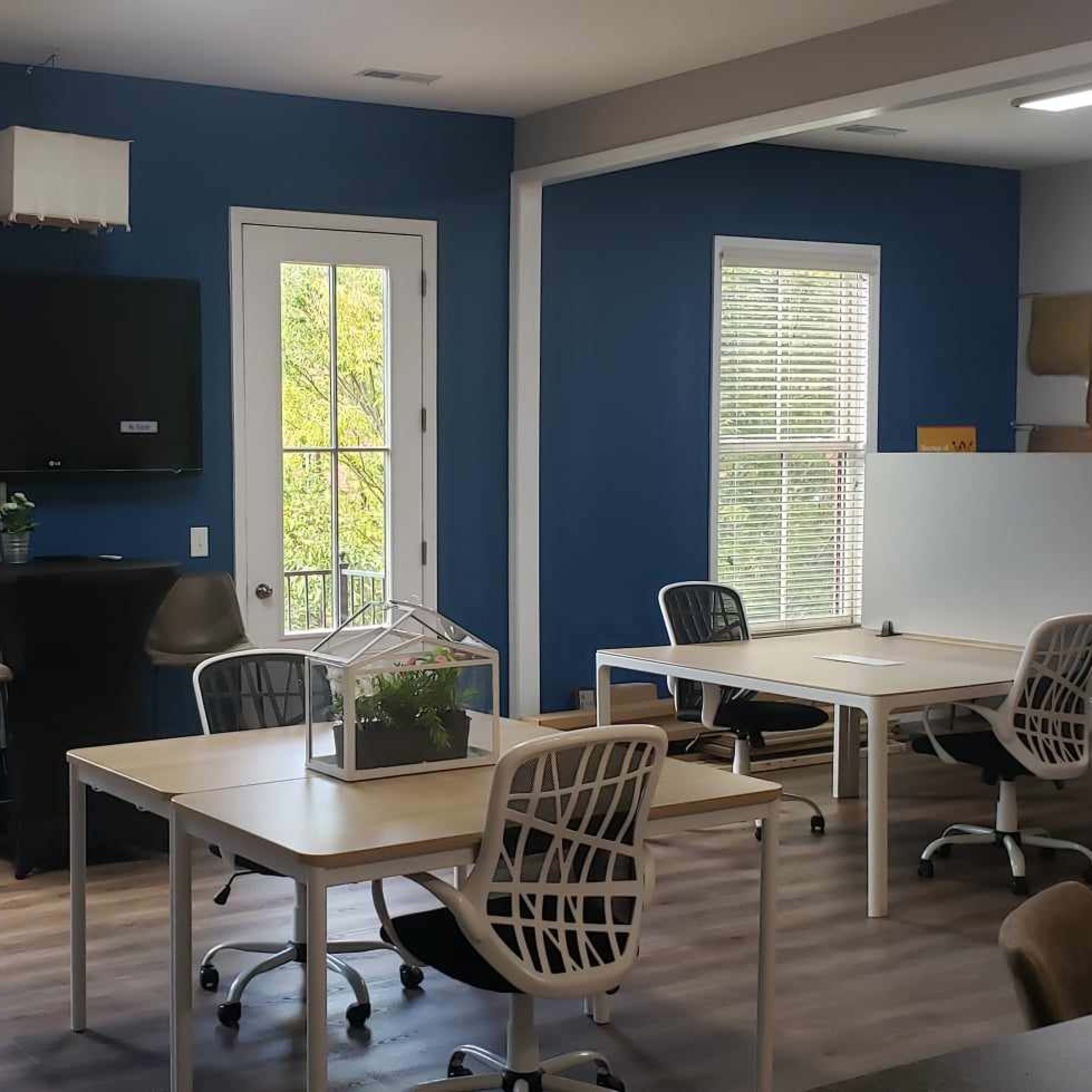 A room with blue walls features several tables and chairs, a television on the wall, and large windows providing natural light.