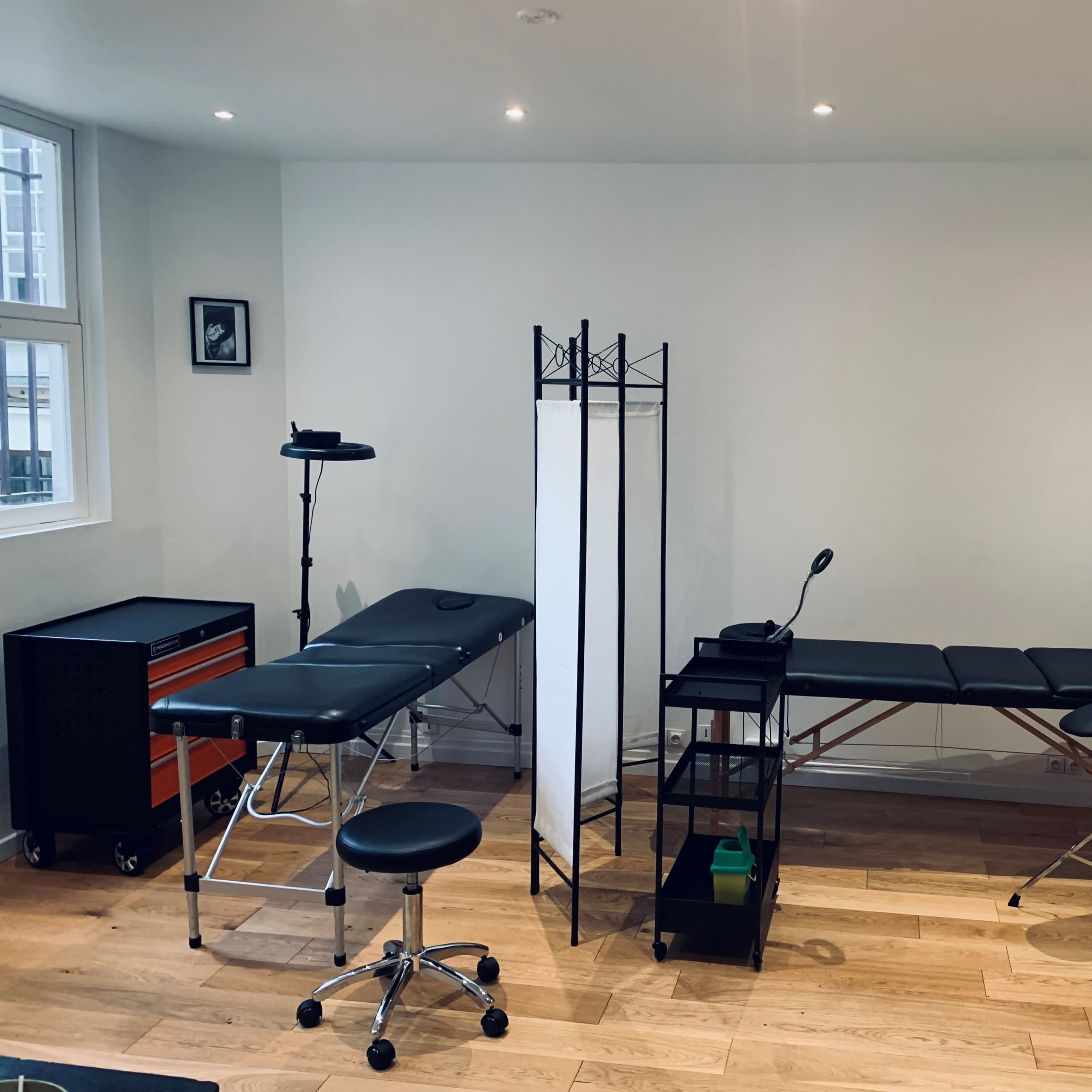A minimalist treatment room featuring two examination tables, a privacy screen, a task light, and a shelving unit against a white wall.