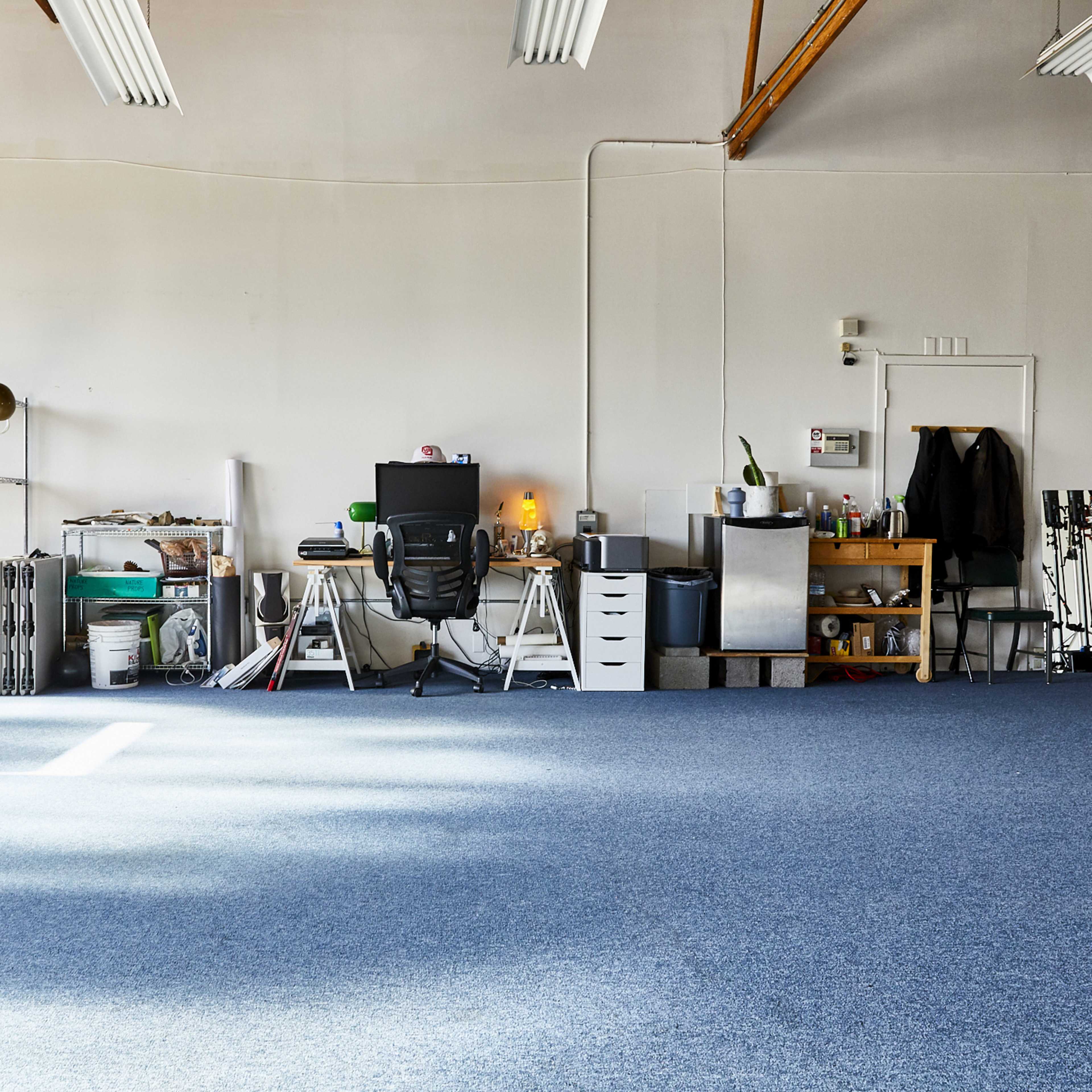 A spacious, empty room with blue carpet, featuring shelves with various items, a desk with a computer and chair, and a storage table against the wall.