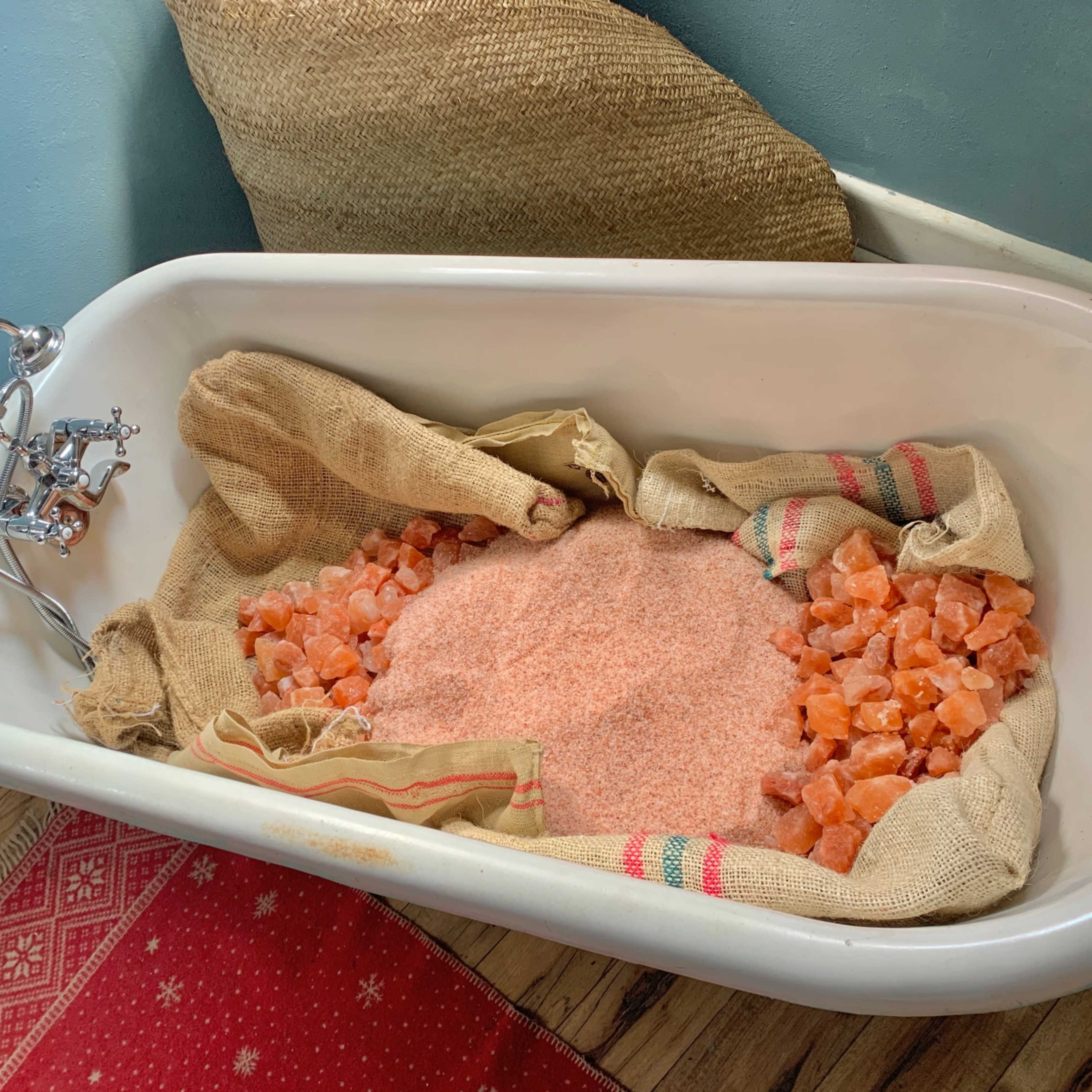 A vintage bathtub is filled with pink Himalayan salt, surrounded by burlap sacks and placed on a wooden floor with a patterned rug.