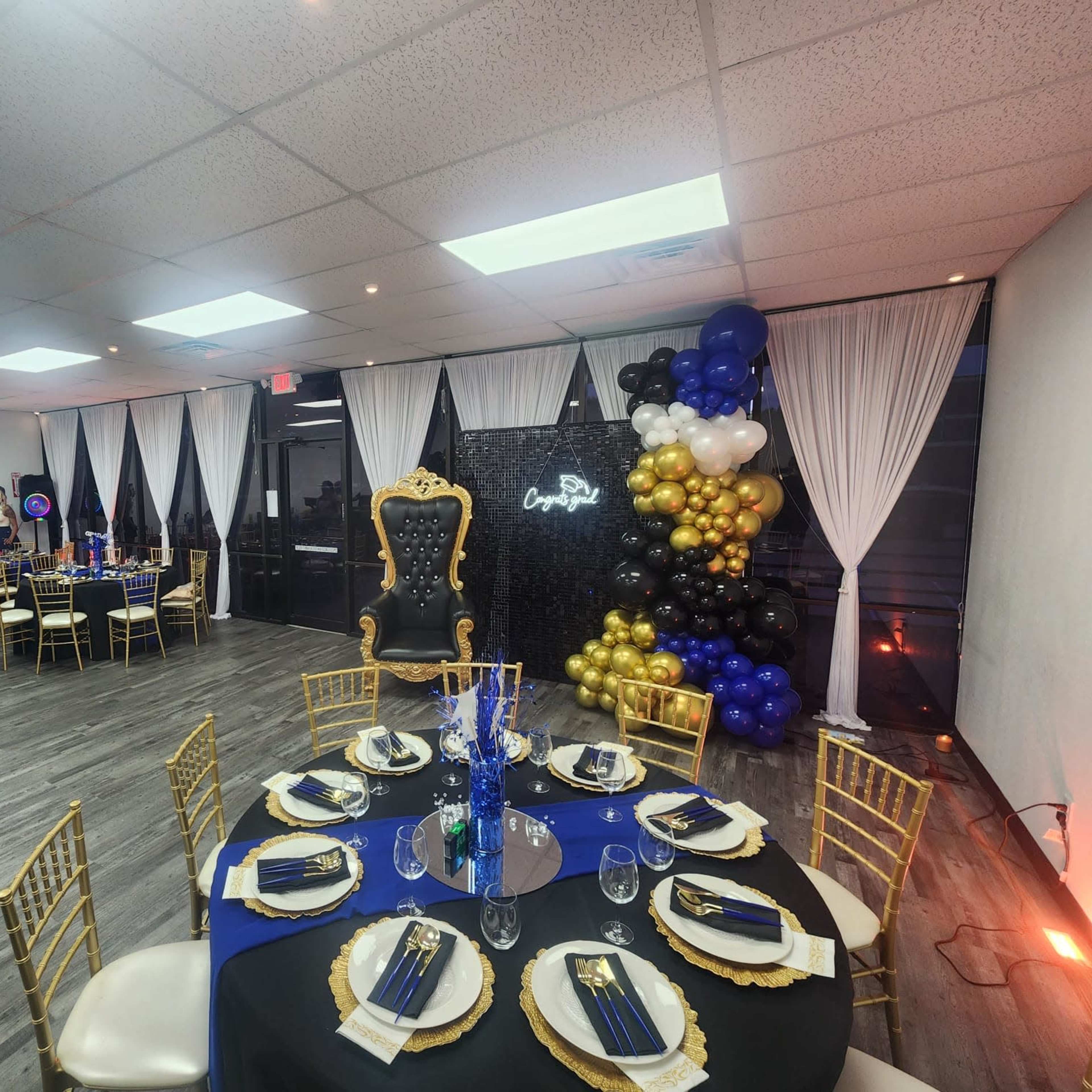 An event space set up for a celebration, featuring elegantly arranged tables with gold chairs, a decorative backdrop, and a colorful balloon arch.