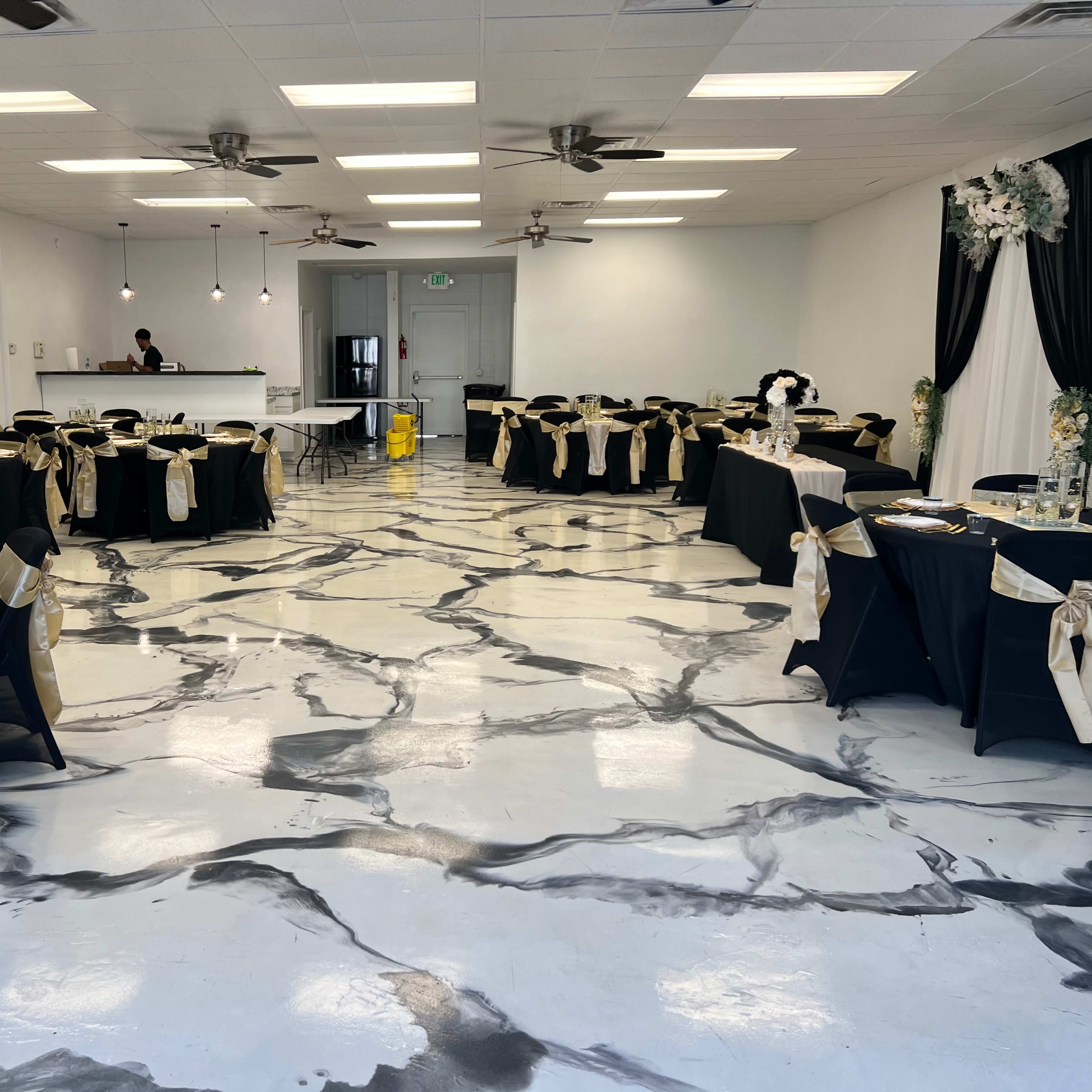 The room features neatly arranged round tables covered with black tablecloths and golden sashes, set against a marbled floor and minimalist decor.