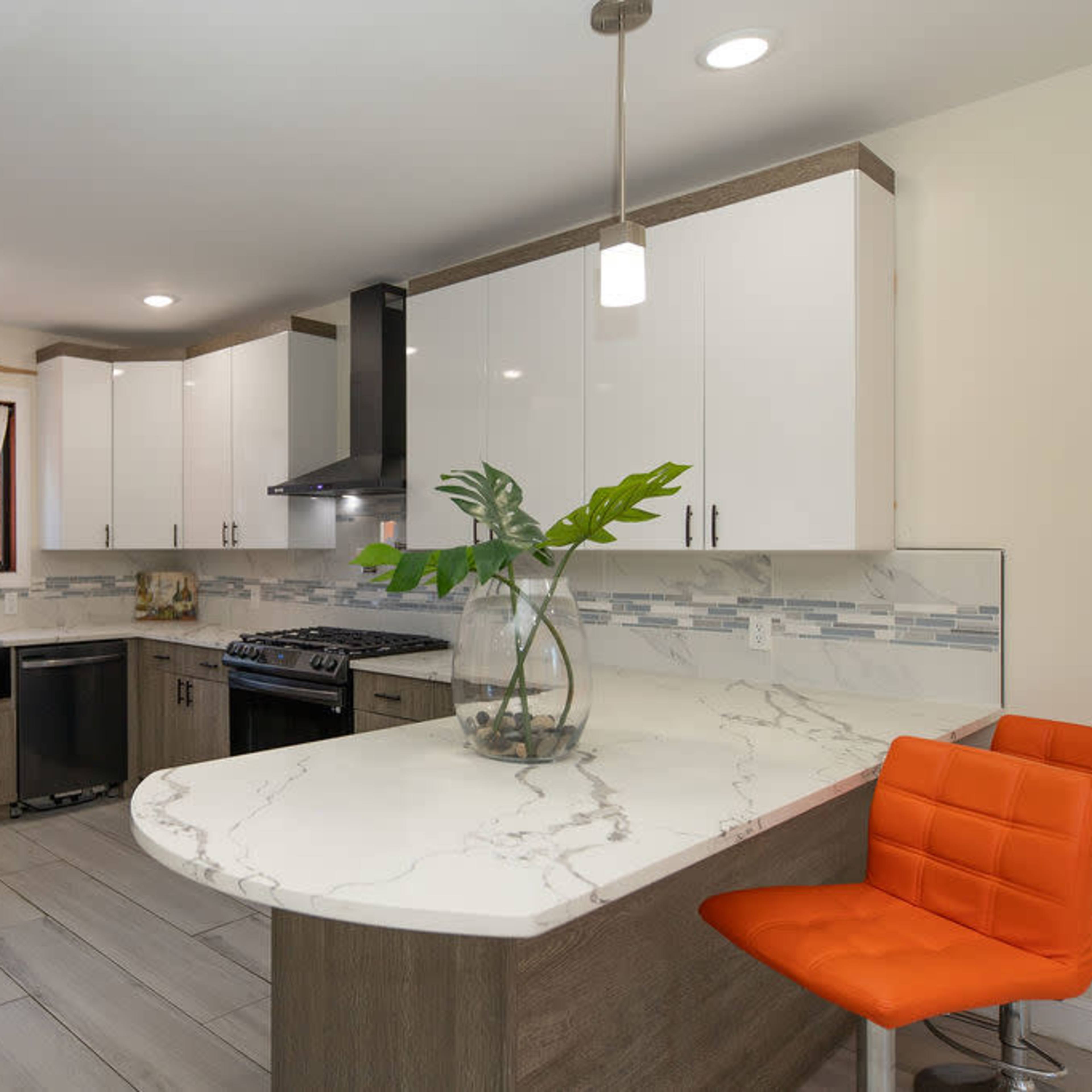 A modern kitchen with white cabinets, a gray countertop, and an orange bar stool at a curved island.