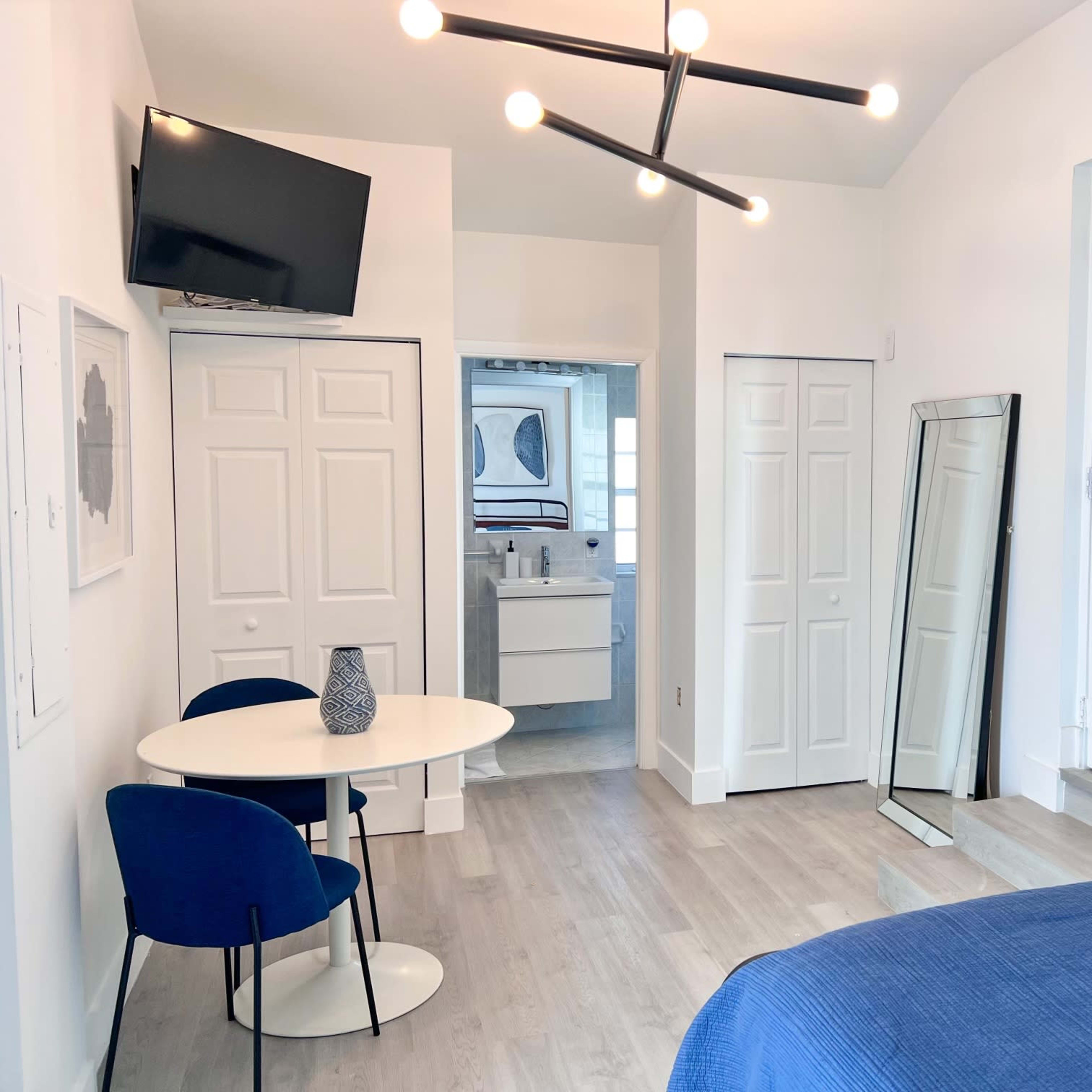 A modern bedroom with a blue bedspread, a small round table and two chairs, a large mirror, and a television mounted on the wall.