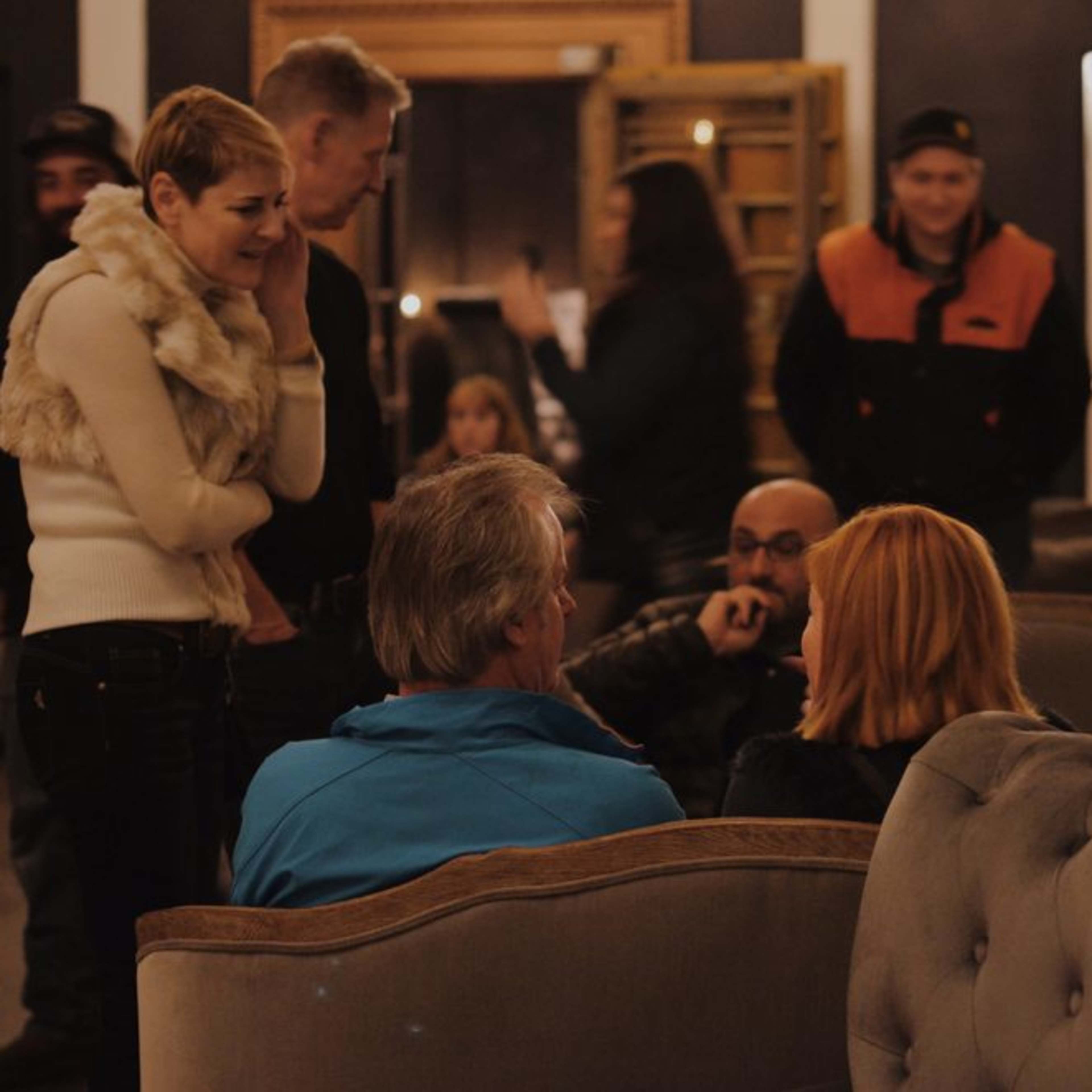 A group of people is gathered in a cozy lounge setting, engaged in conversation, with some seated on plush sofas and others standing nearby.