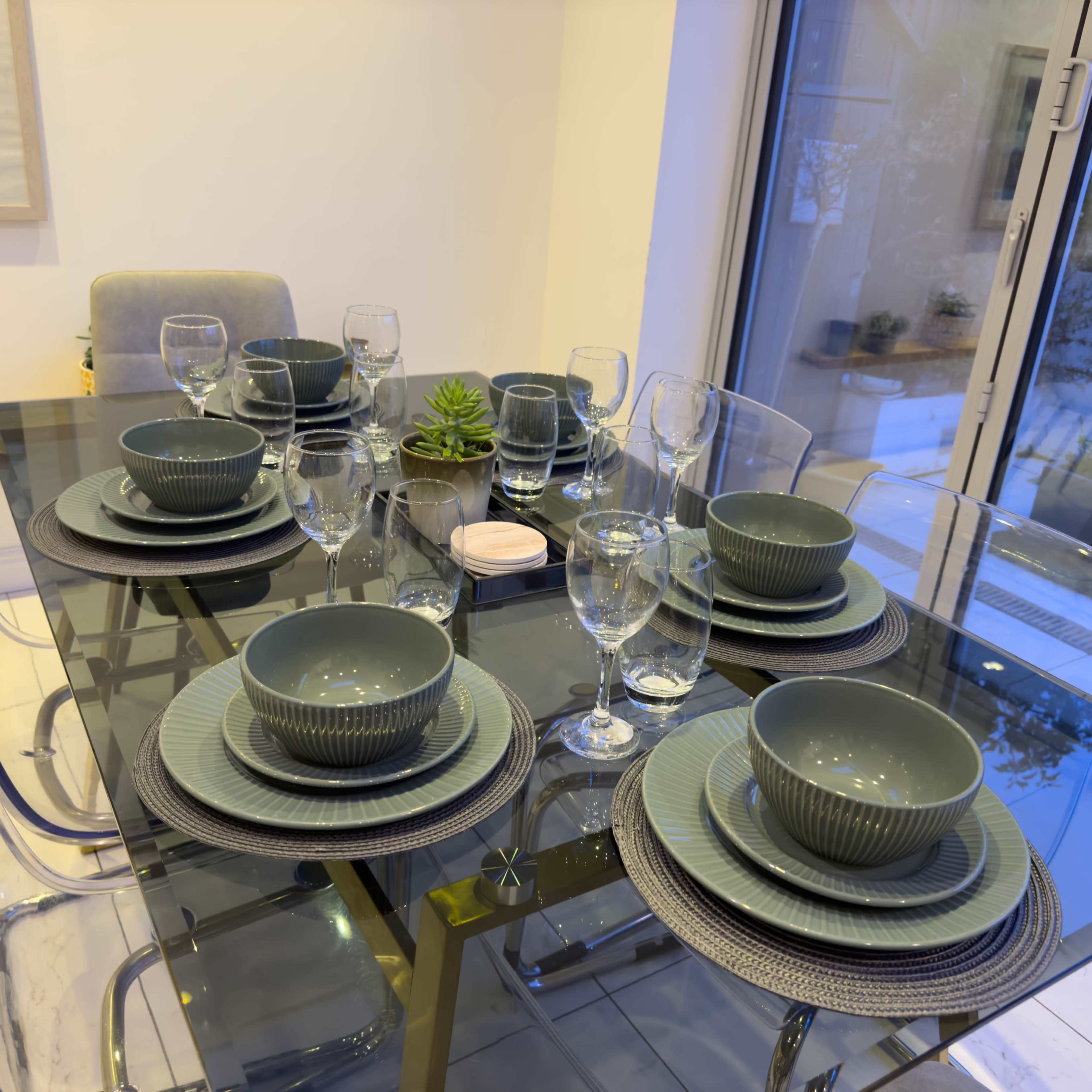 A dining table is set with green dishware, glassware, and decorative elements arranged neatly on a glass surface.