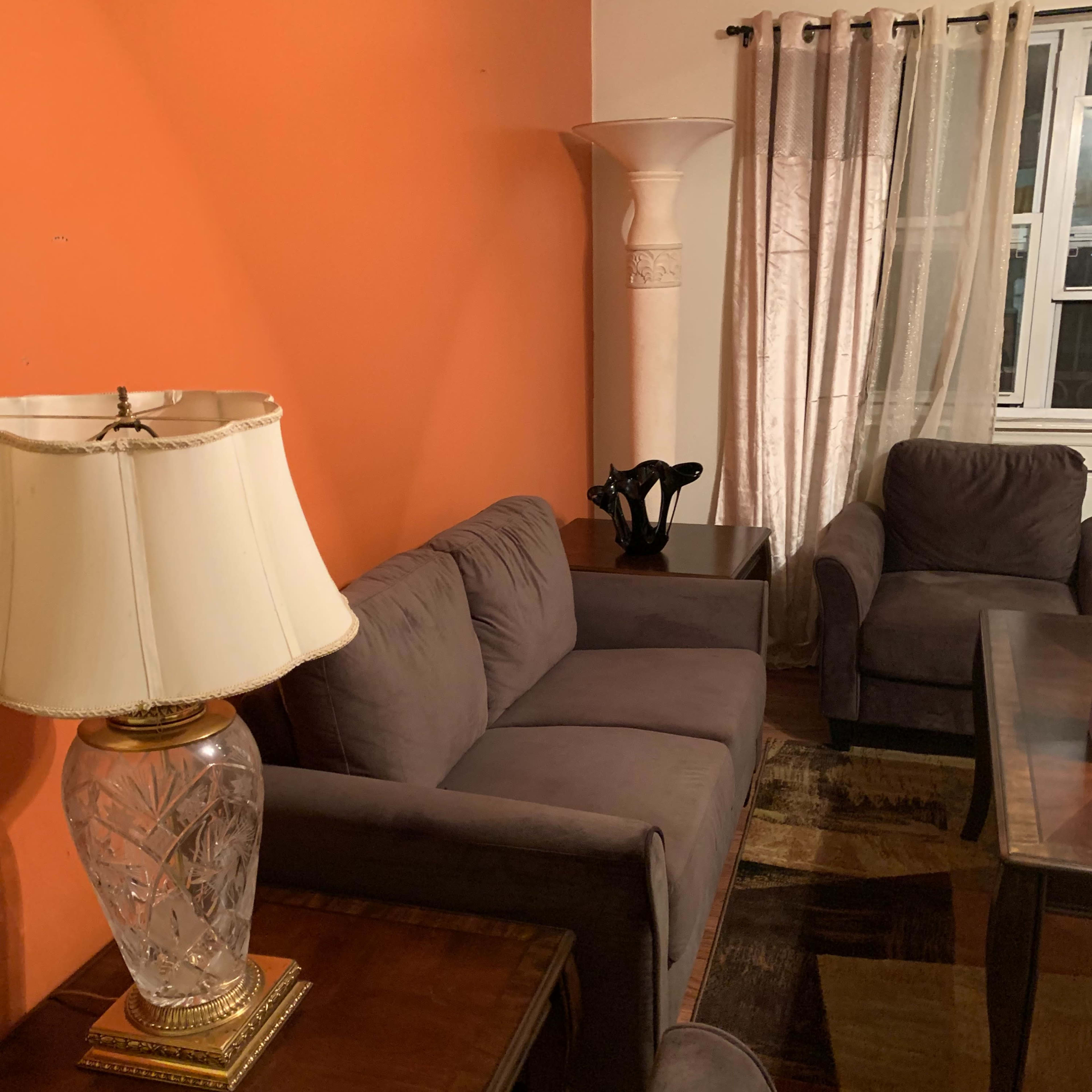 A living room with a gray sofa and armchairs, a glass lamp on a wooden table, and an orange wall with curtains next to a window.