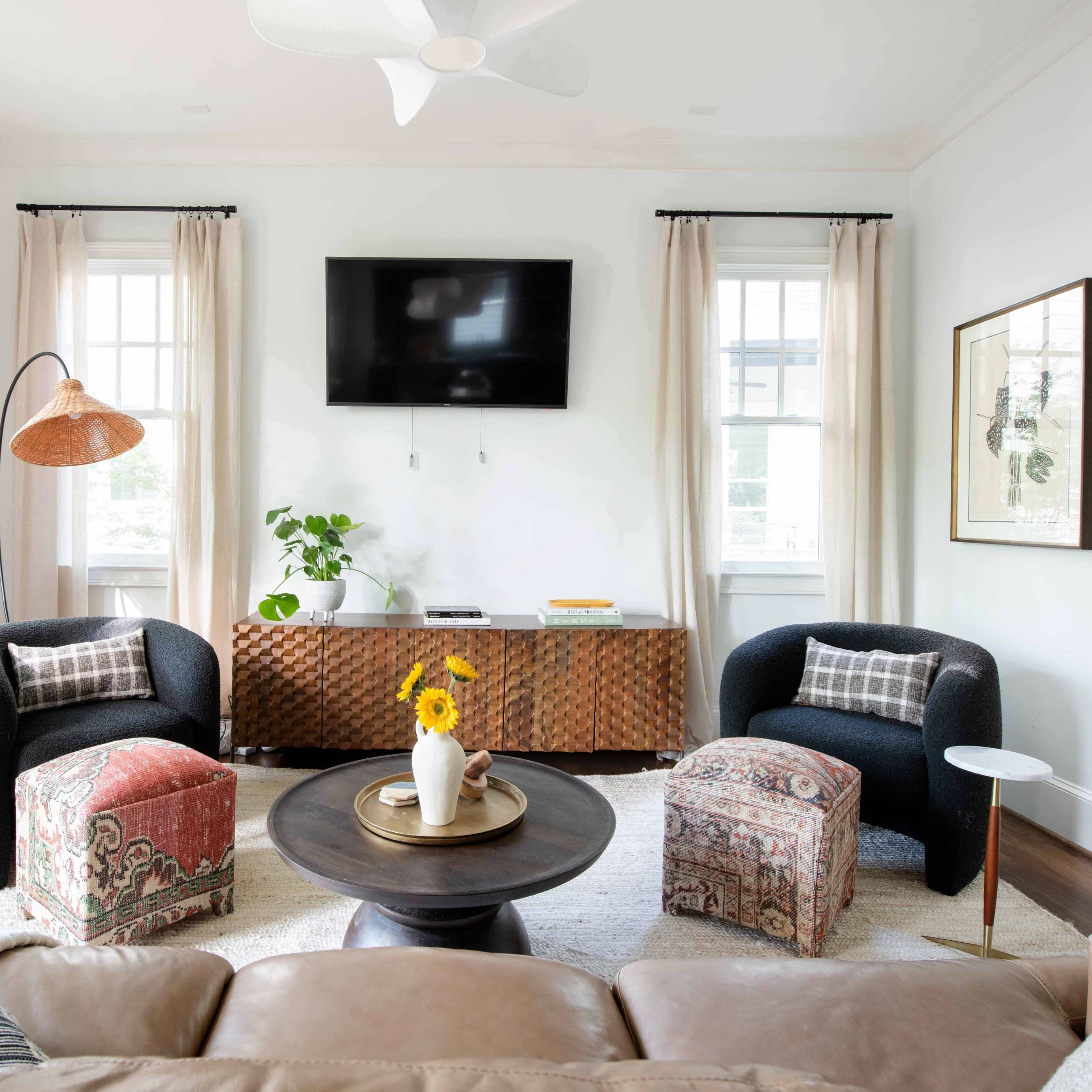 The living room features two black armchairs, a round coffee table, and a wooden sideboard with art on the walls, illuminated by natural light through large windows.