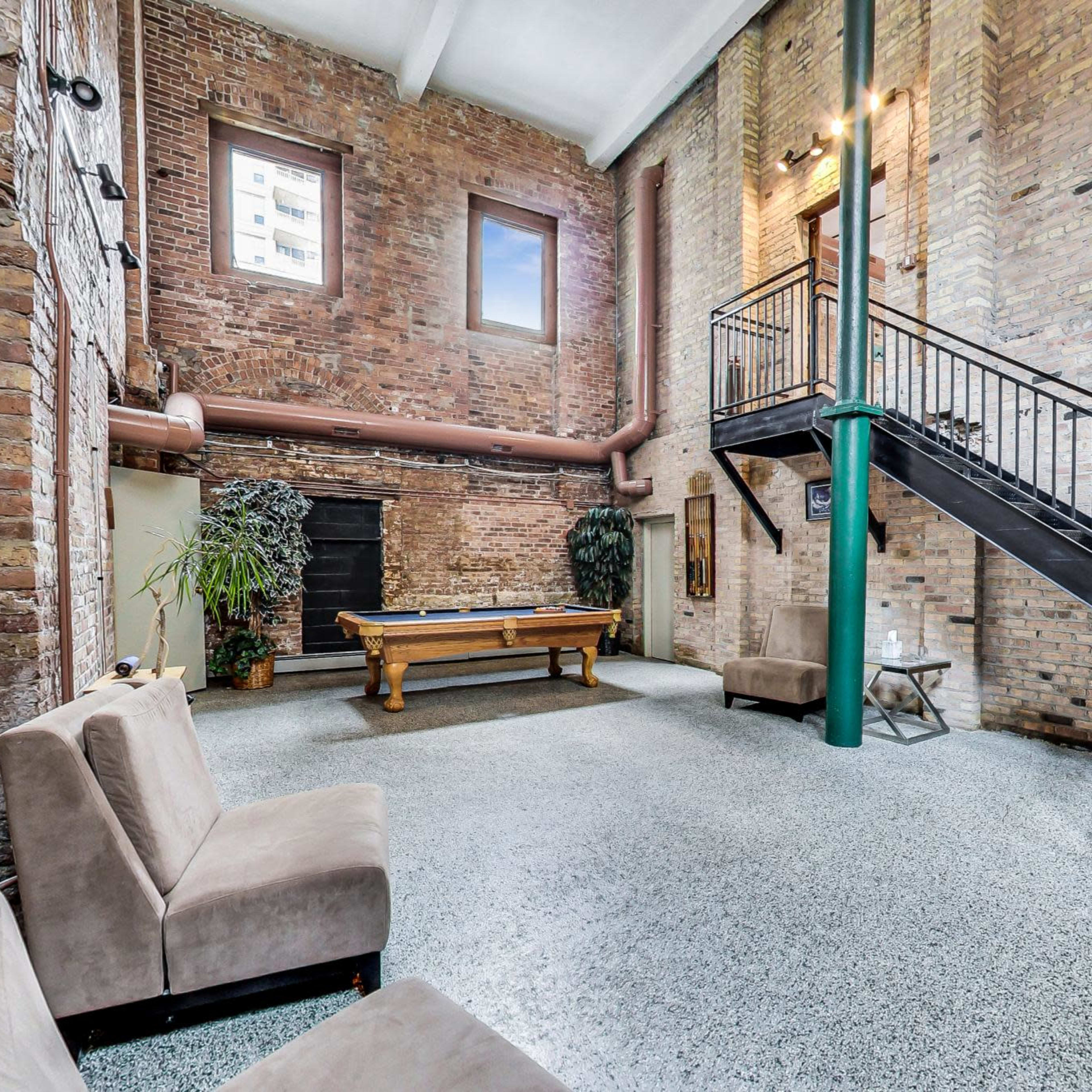 An industrial-style room featuring exposed brick walls, a staircase, and a pool table, with seating arranged to the side.