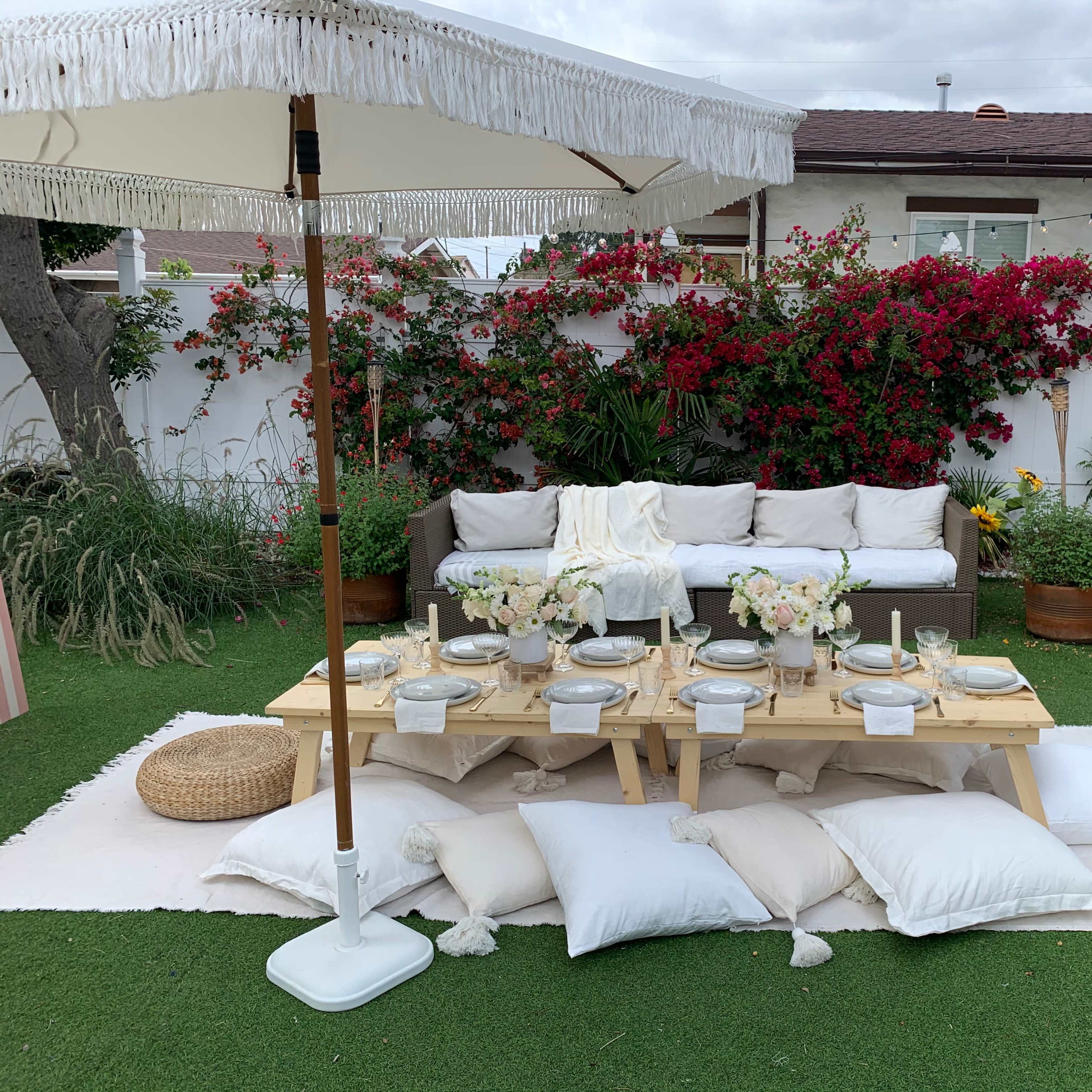 A garden seating area features a low wooden table adorned with floral arrangements and surrounded by white pillows, with a large umbrella providing shade.