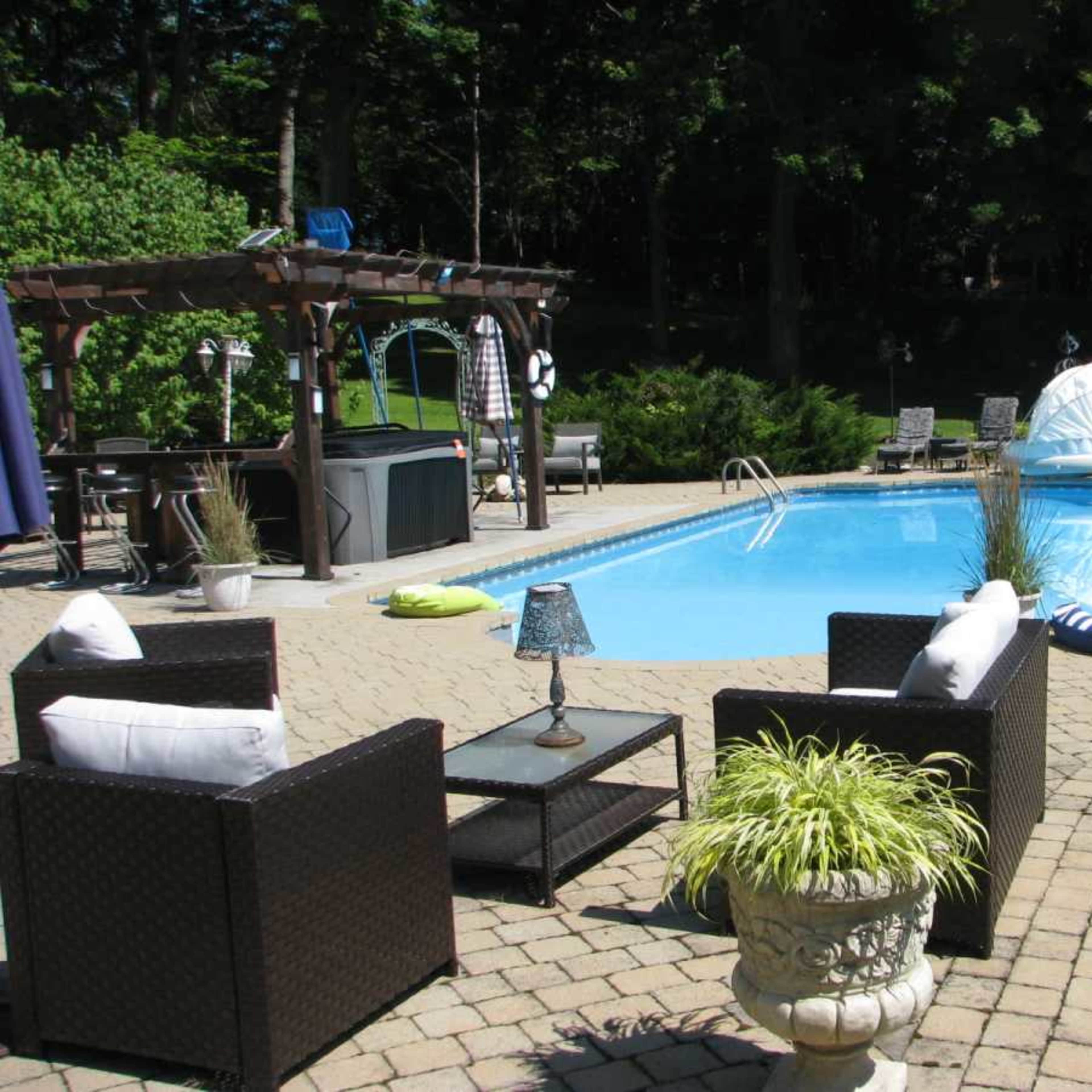 A backyard scene featuring a rectangular swimming pool surrounded by decorative landscaping, seating areas with wicker furniture, and a small shed in the background.