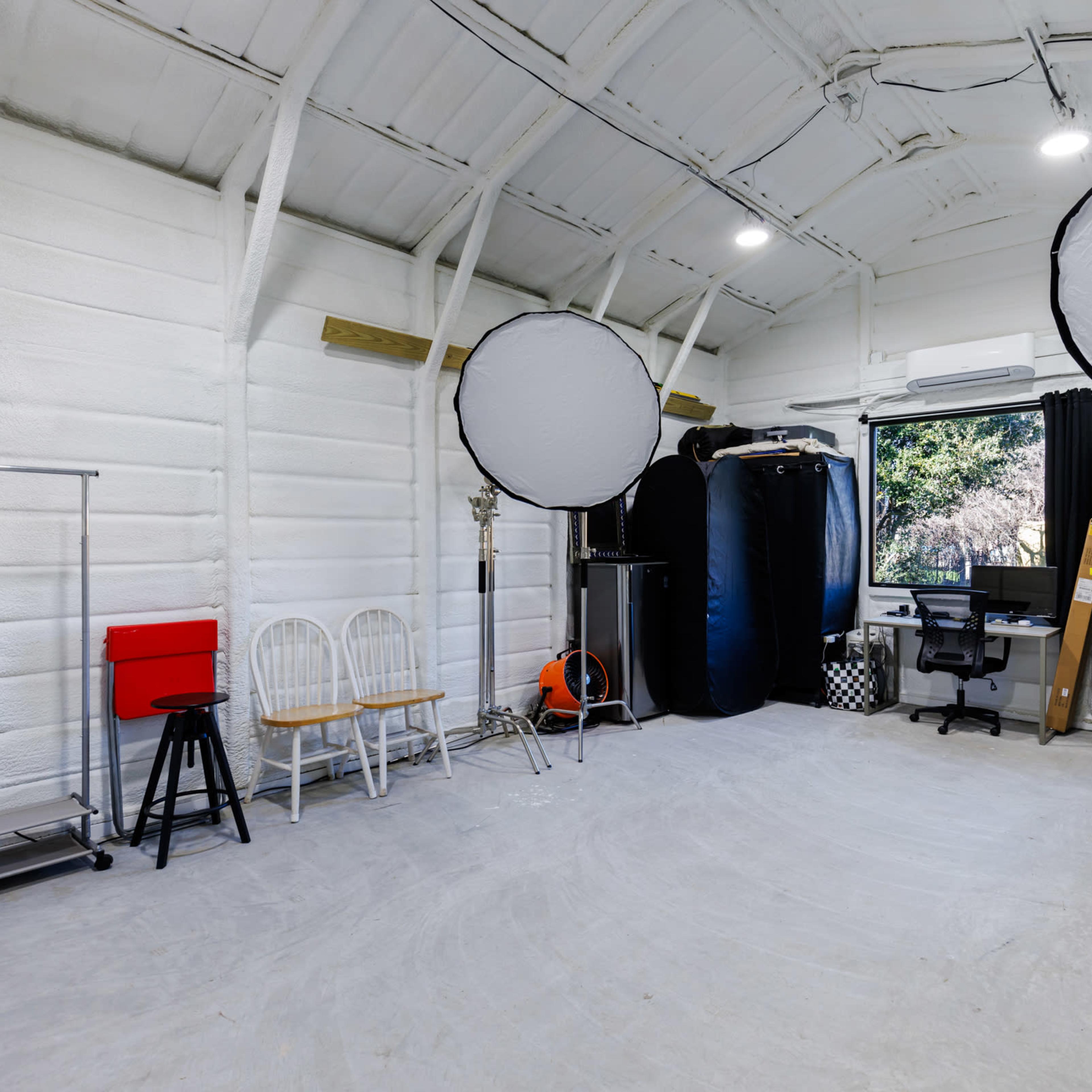 A spacious studio with white walls, two large light diffusers, a desk and chair, and various equipment along the sides.