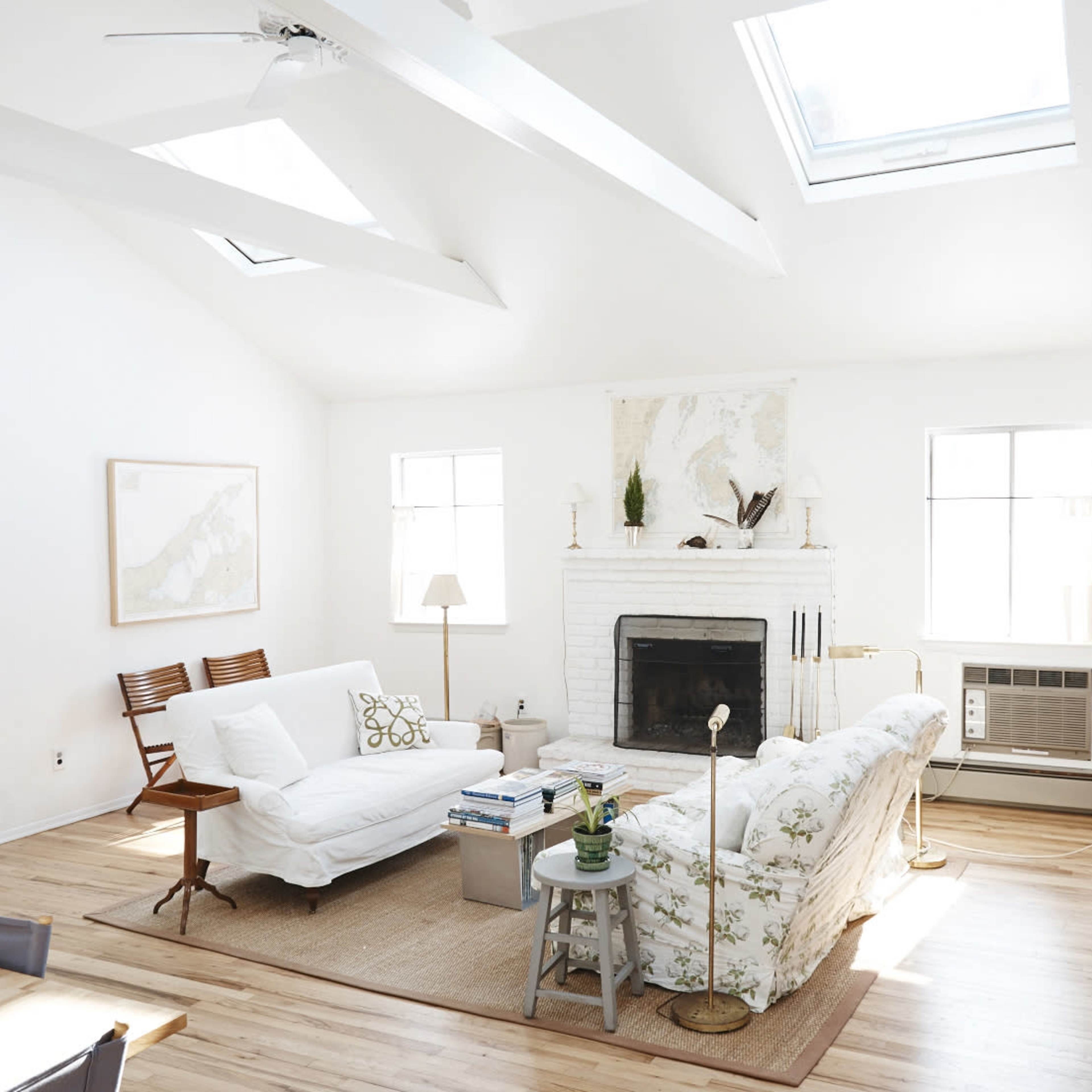 A bright, airy living room featuring a white sofa, beige armchair, and a fireplace, with large windows and skylights illuminating the space.