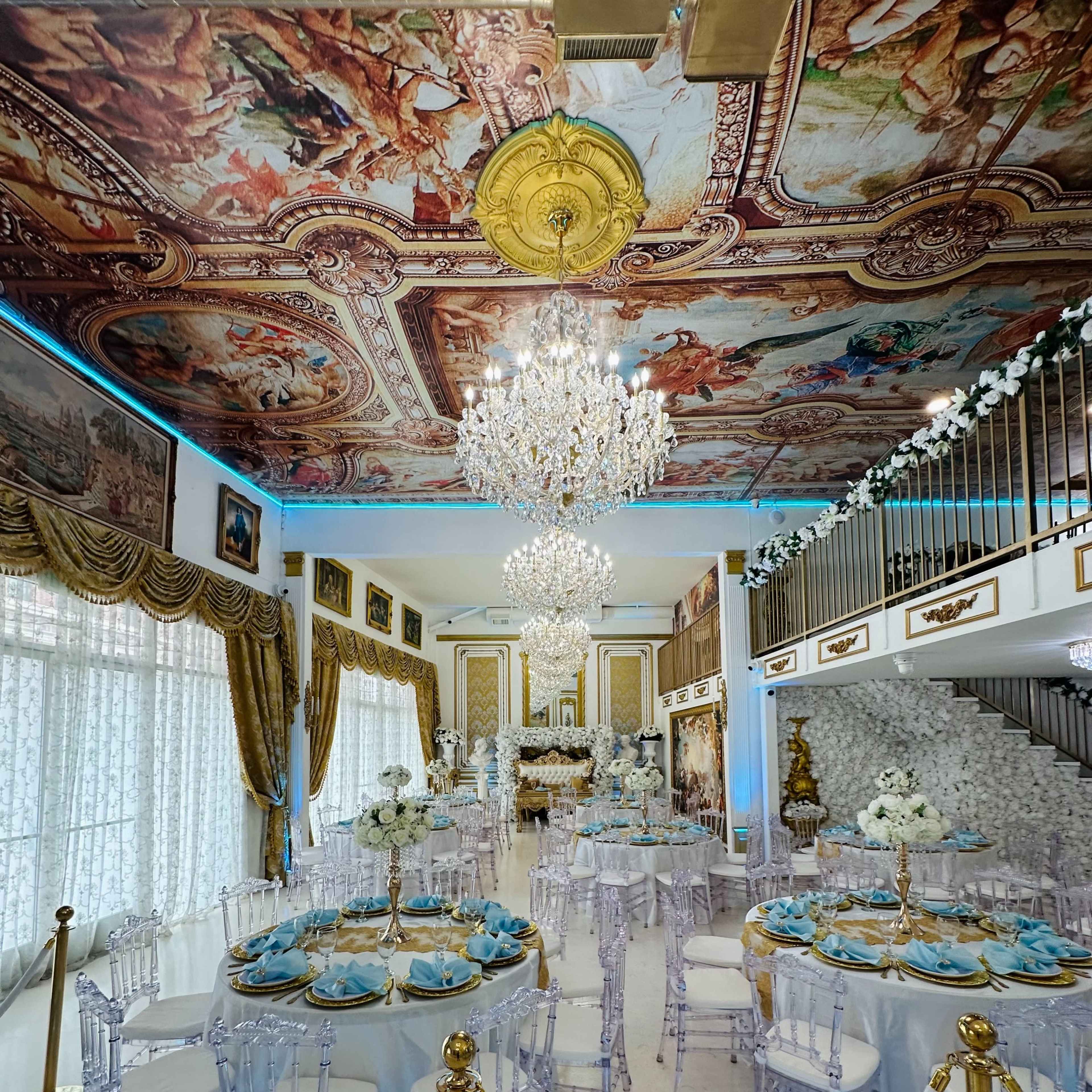 The elegant banquet hall features ornate ceiling artwork, a chandelier, and tables set with blue and white decor.