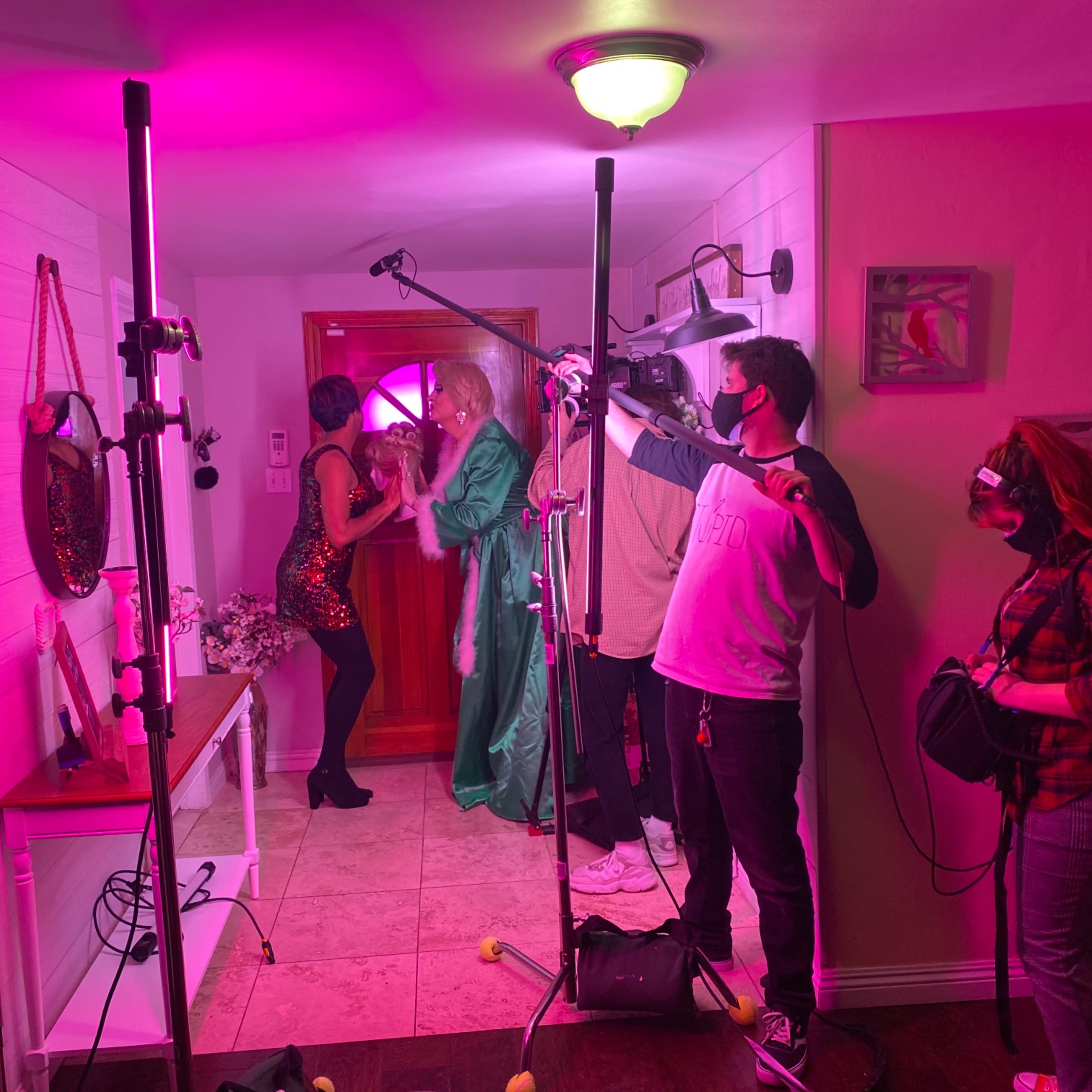A film crew sets up equipment in a brightly lit room while two actors perform near a doorway.