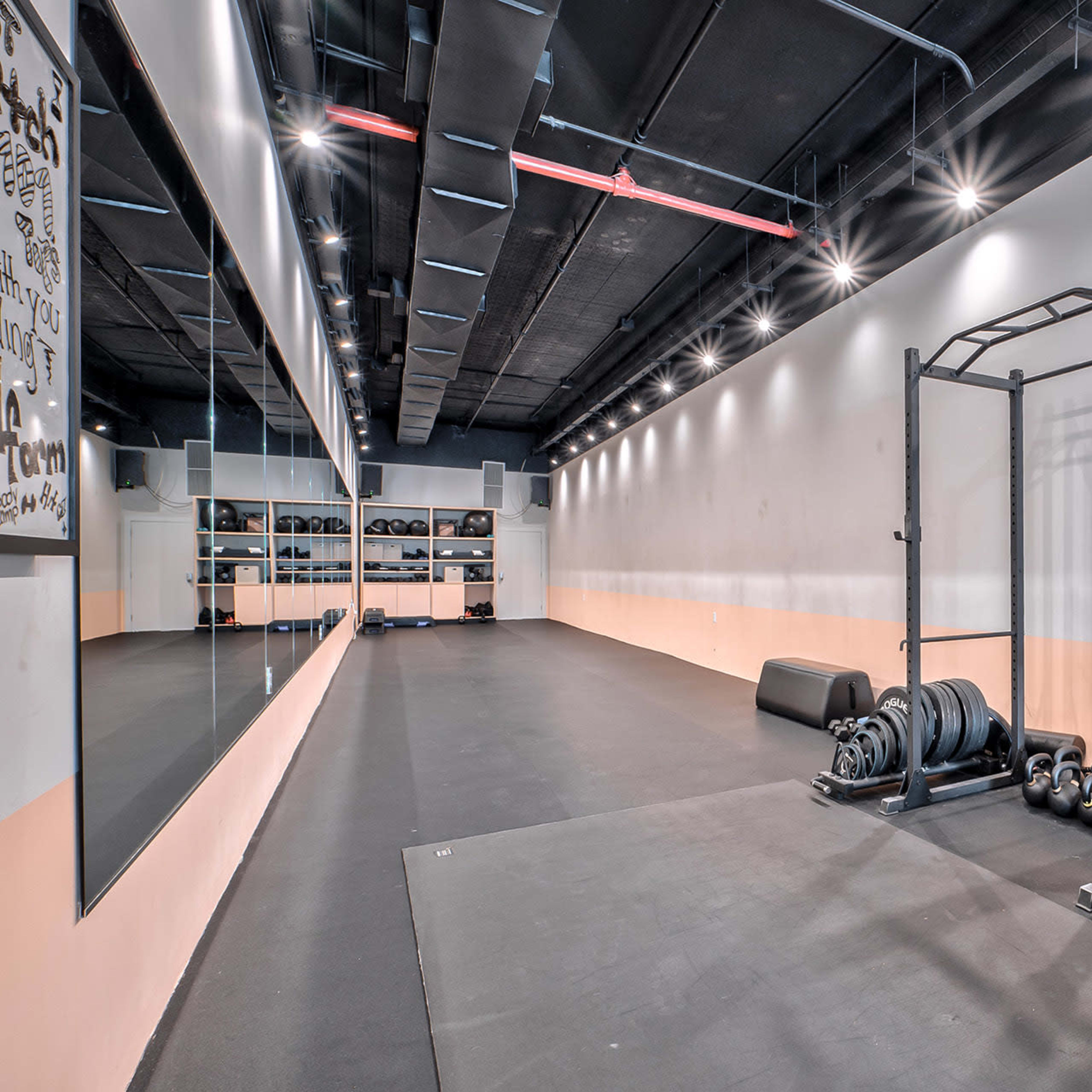 A spacious gym interior with a black rubber floor, weights stored on shelves, a squat rack, and large mirrors along the wall.