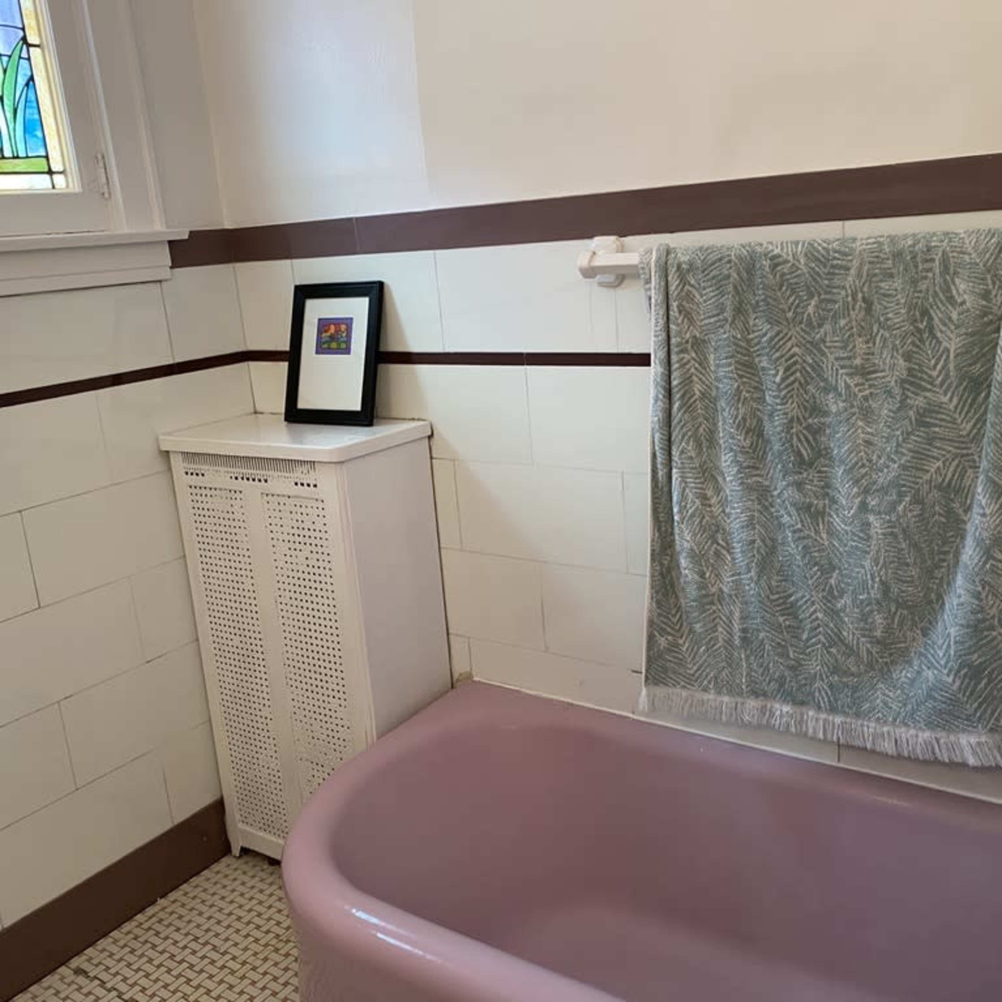 A pink bathtub is positioned in a bathroom with white tiled walls, a green patterned towel hanging on a rack, and a small framed picture on a shelf.