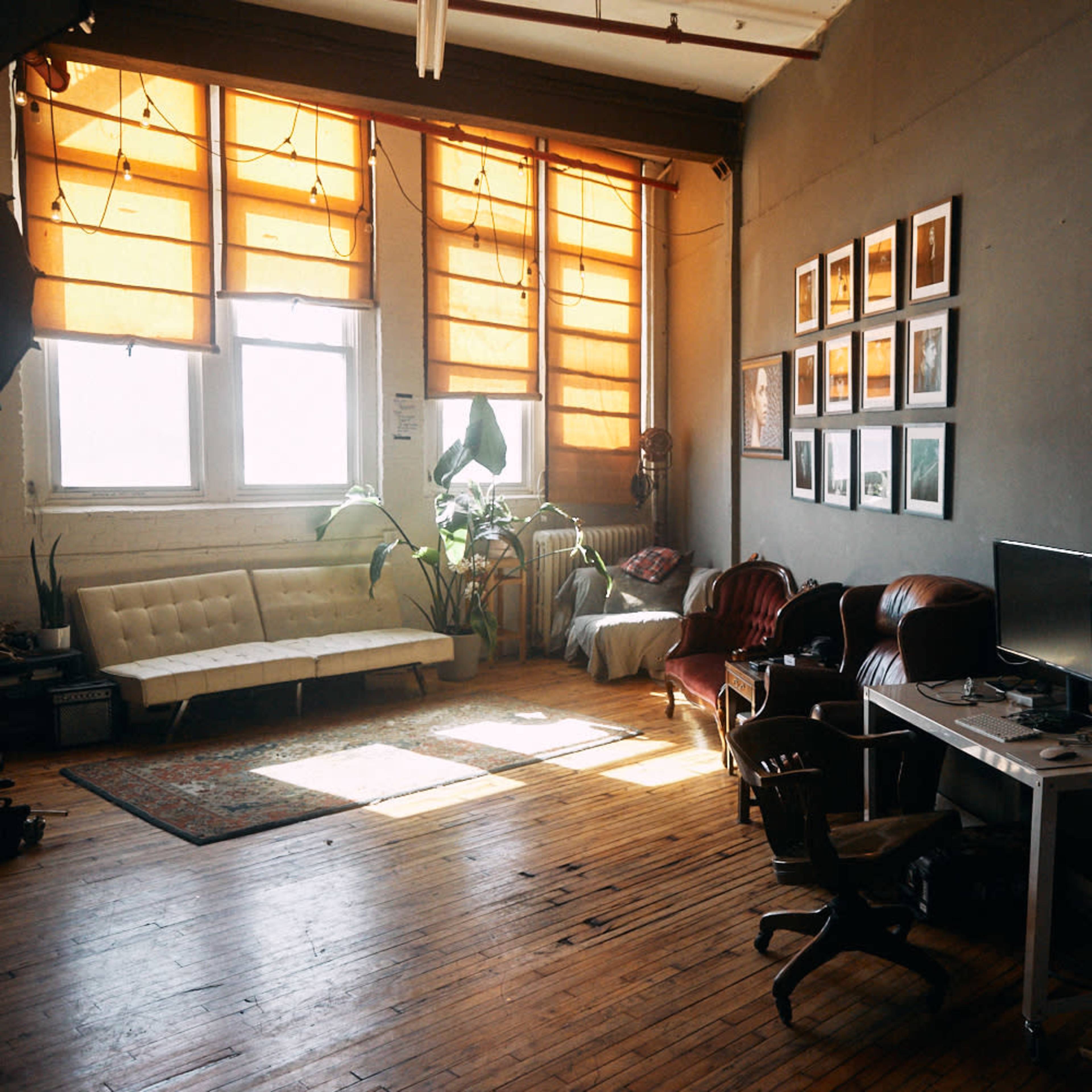 A spacious, sunlit room with wooden floors, featuring a sofa, armchairs, a coffee table, and a desk with a computer, along with several framed photos on the wall.