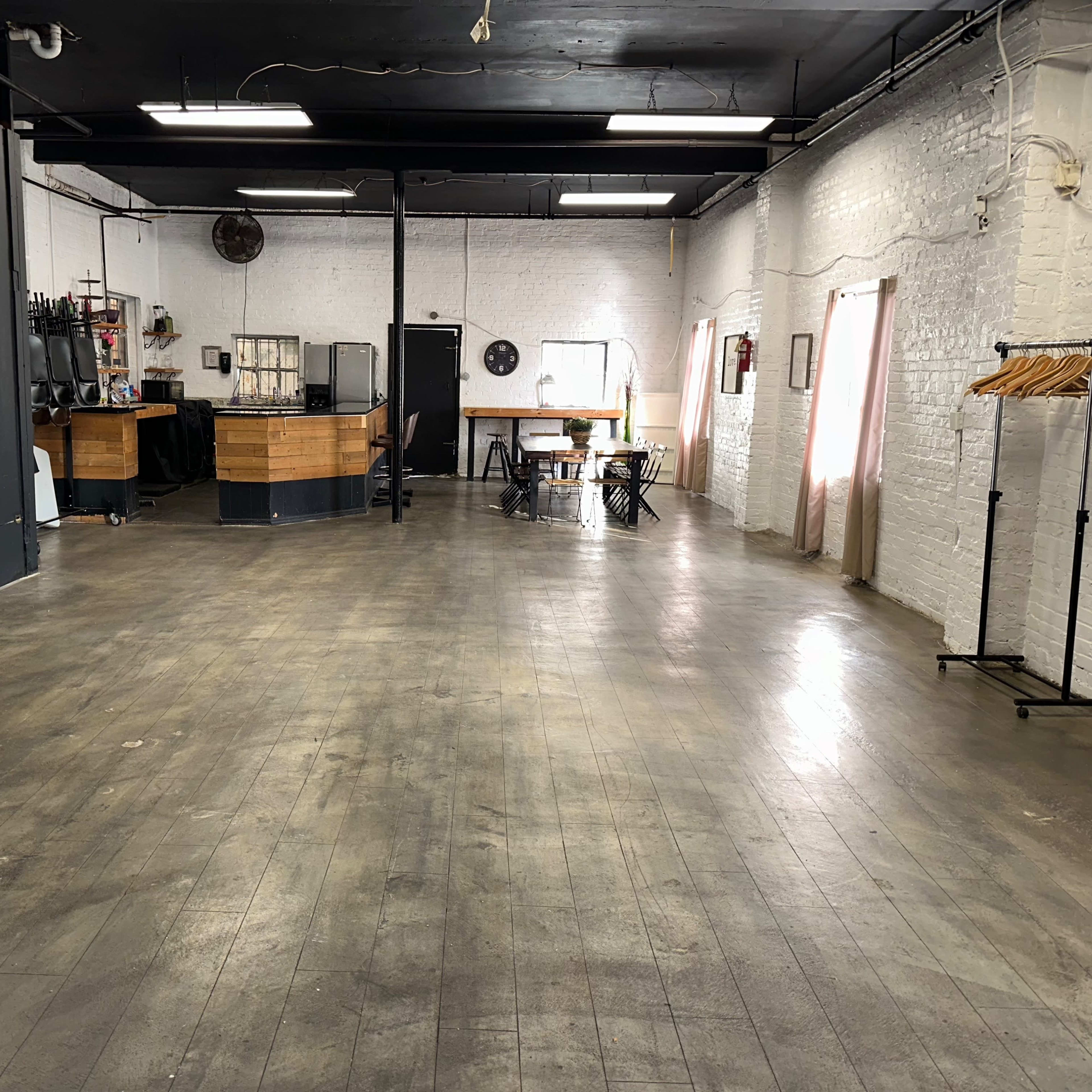 An empty indoor space with wooden flooring, exposed brick walls, and a small bar area in the background.
